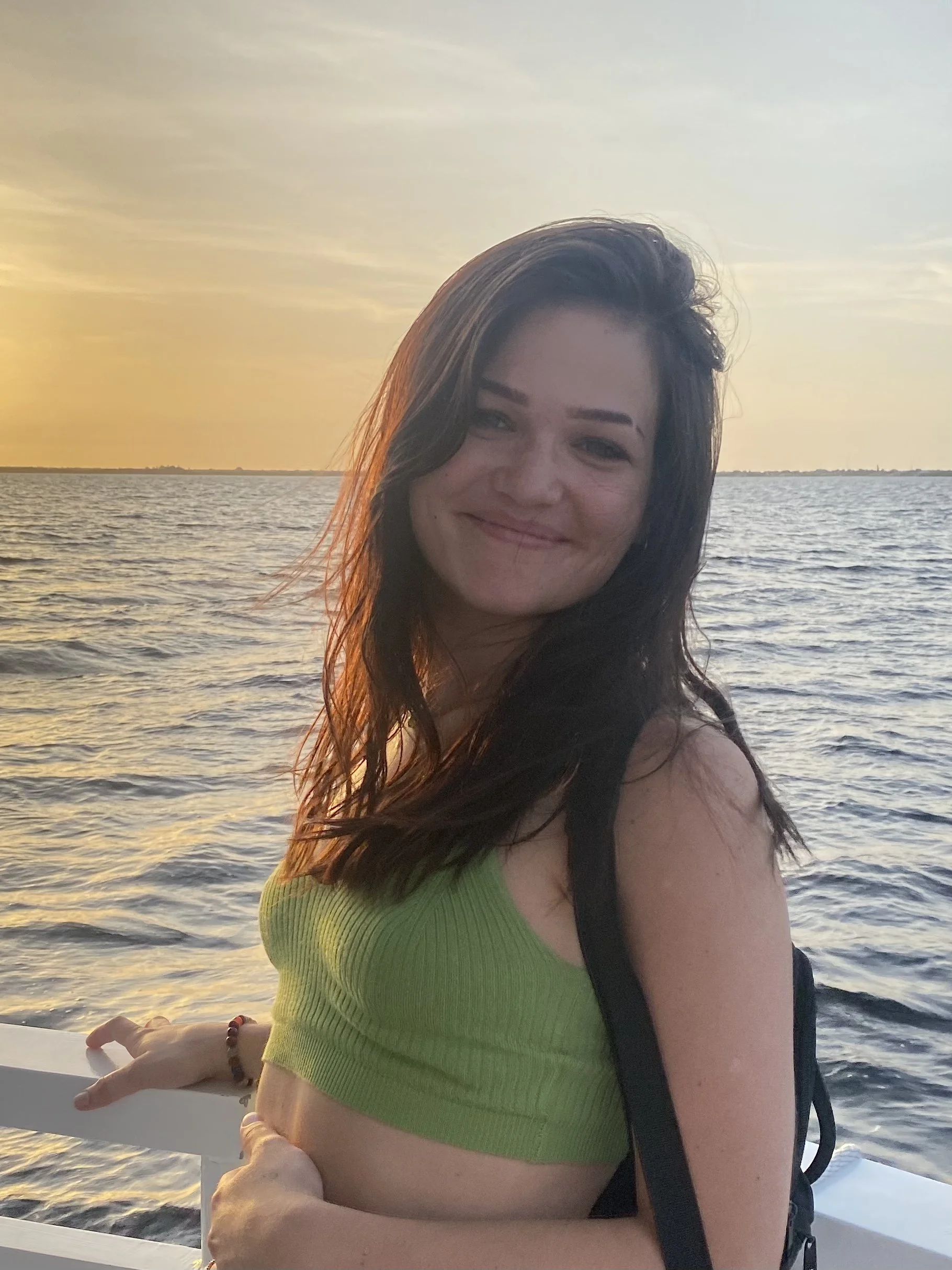 A smiling woman with long brown hair wearing a green crop top standing on a boat at sunset.