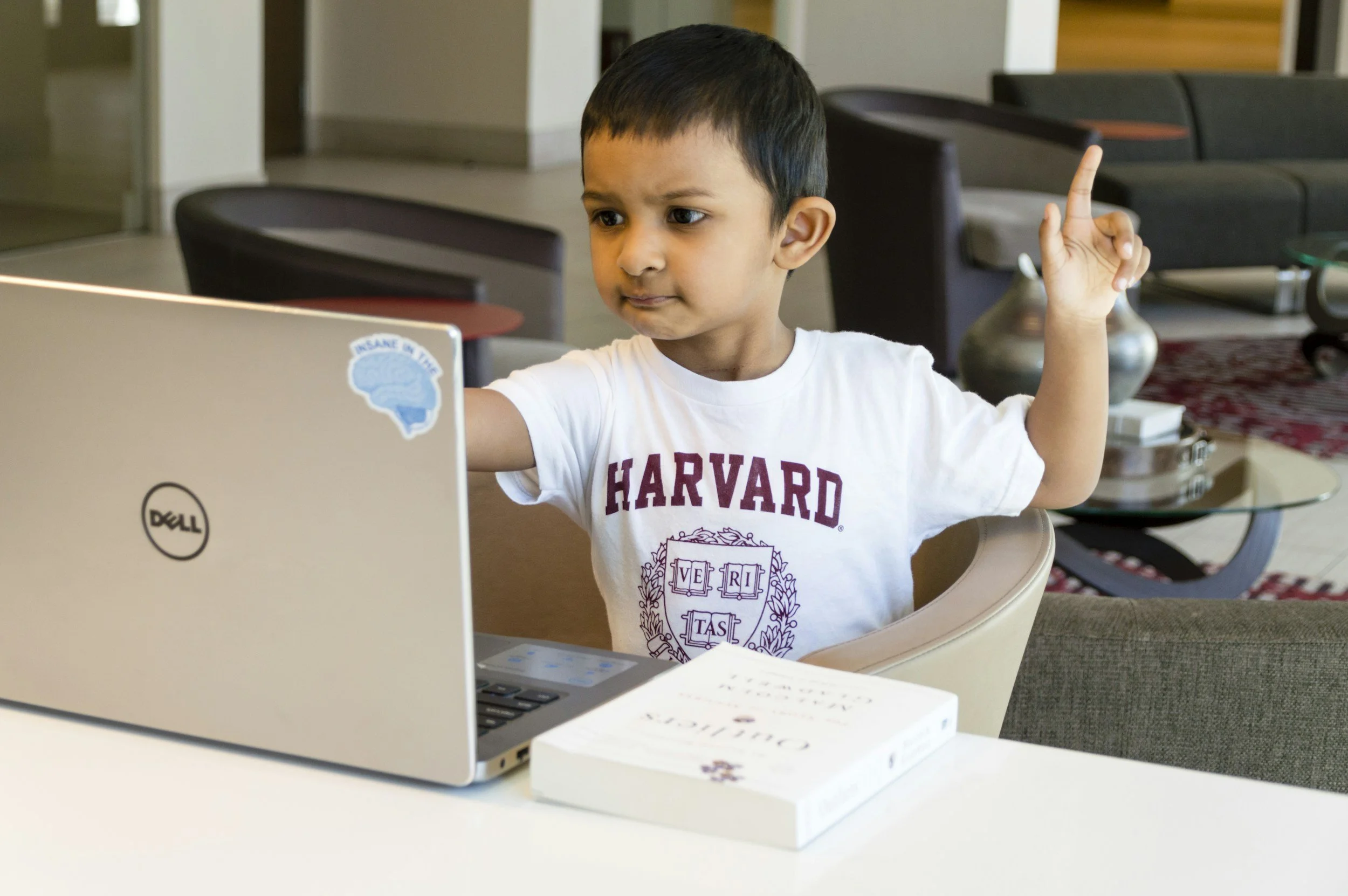 Young boy with short black hair, wearing a white Harvard shirt, sitting at a table and raising his right hand, in a room with modern furnishings, looking at a silver Dell laptop with a sticker on the corner.