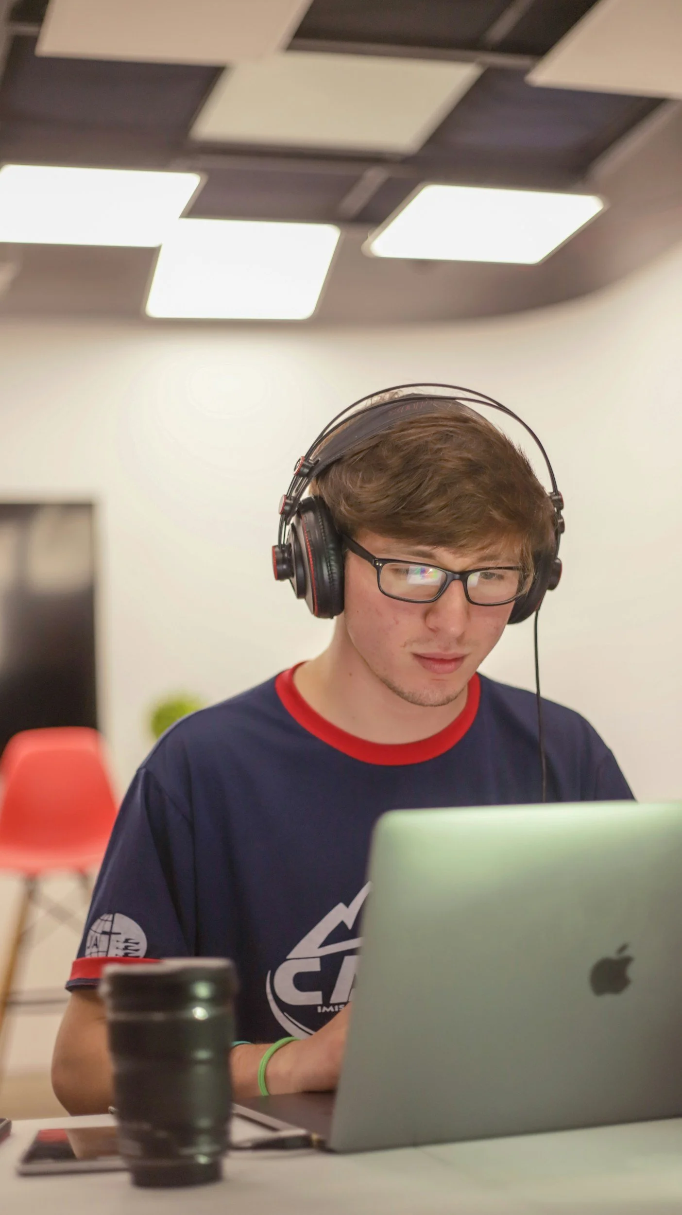 A young man with glasses and headphones working on a laptop at a desk. In the background, there are red chairs and a plain white wall, with ceiling lights overhead.