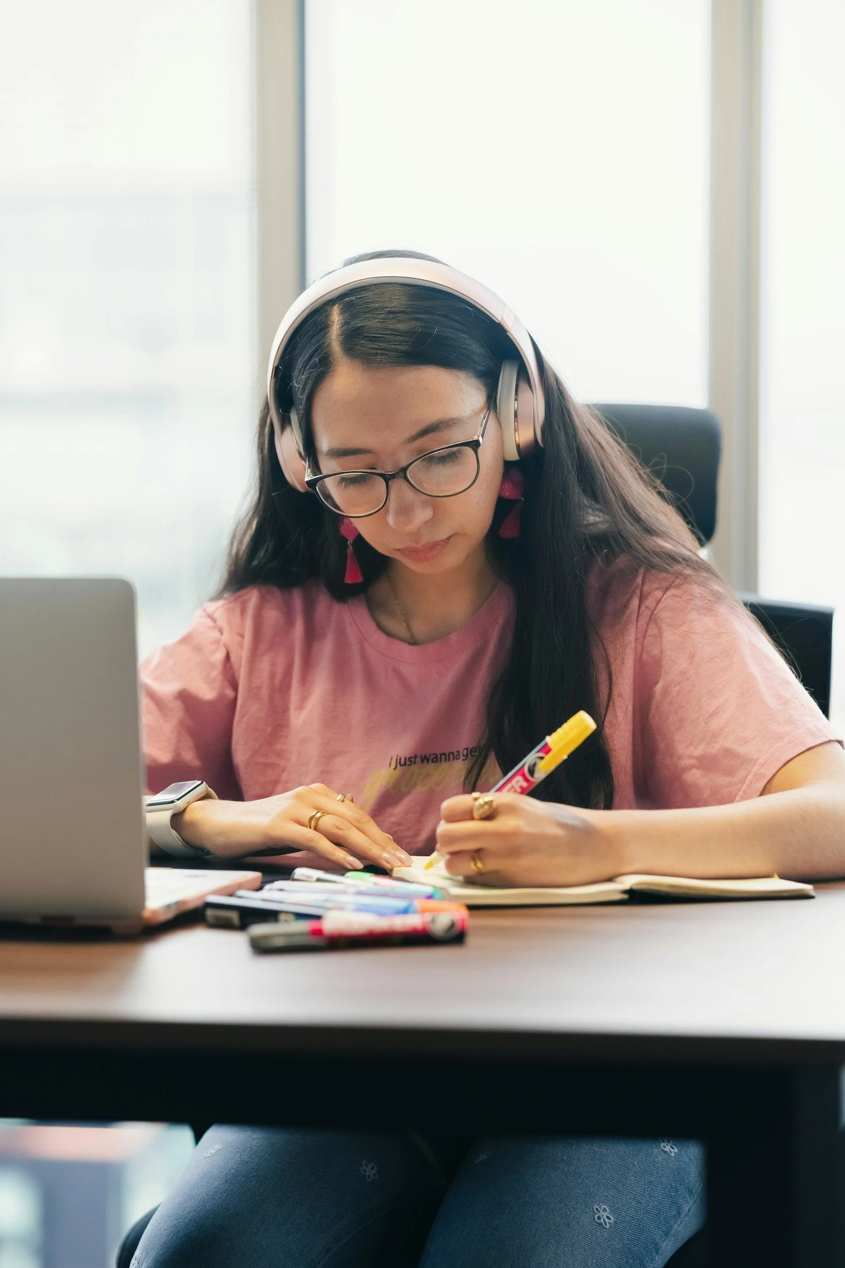 A young woman with long dark hair, glasses, and pink earrings, seated at a desk with a laptop, writing with a yellow marker on paper. She wears a pink T-shirt, and on her left wrist is a smartwatch. In front of her are various colorful markers. She is in a bright room with large windows.