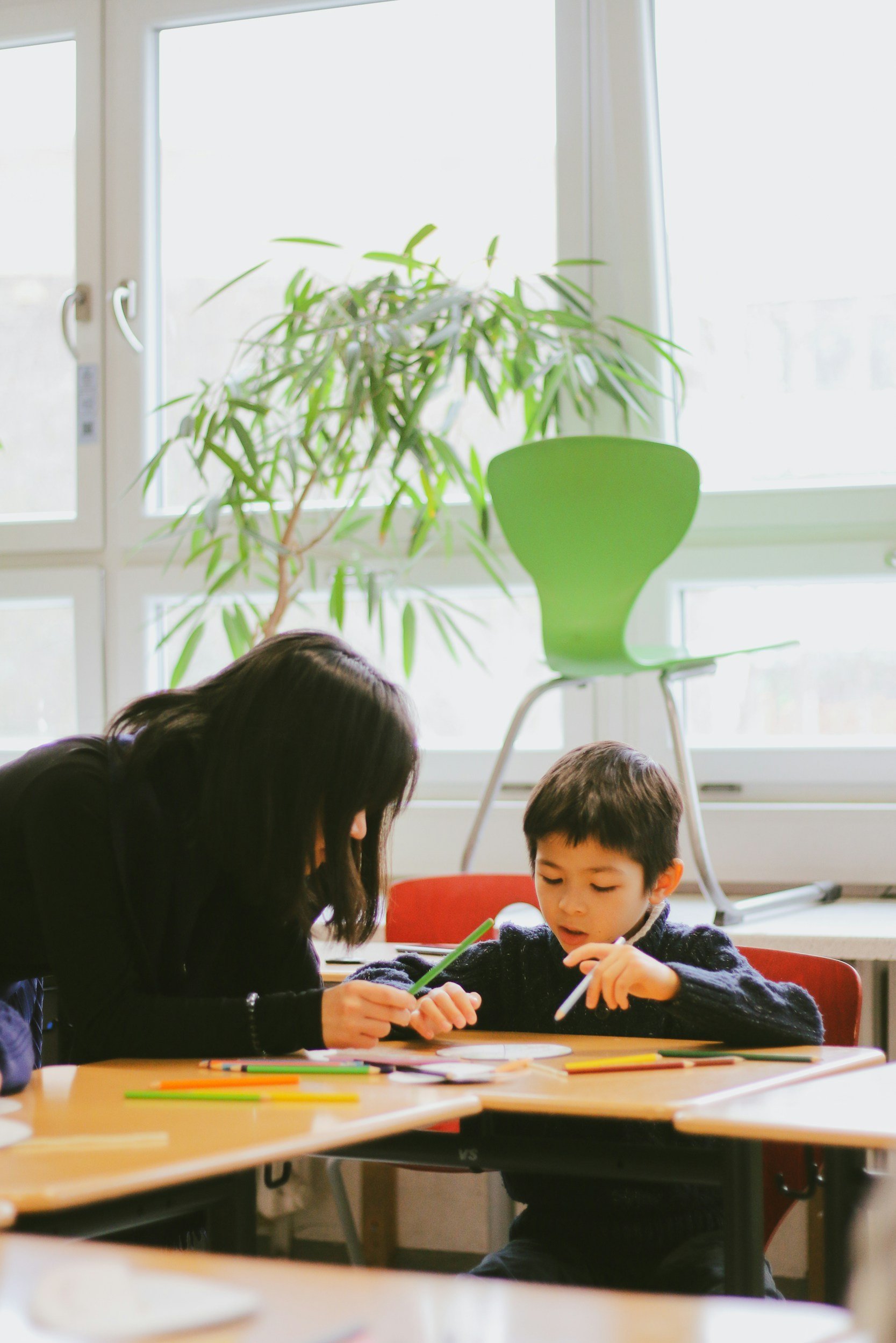A woman and a young boy sitting at a table indoors, engaged in an activity with colorful markers. In the background, there is a large window with bright natural light, a green office chair, and a potted plant near the window.