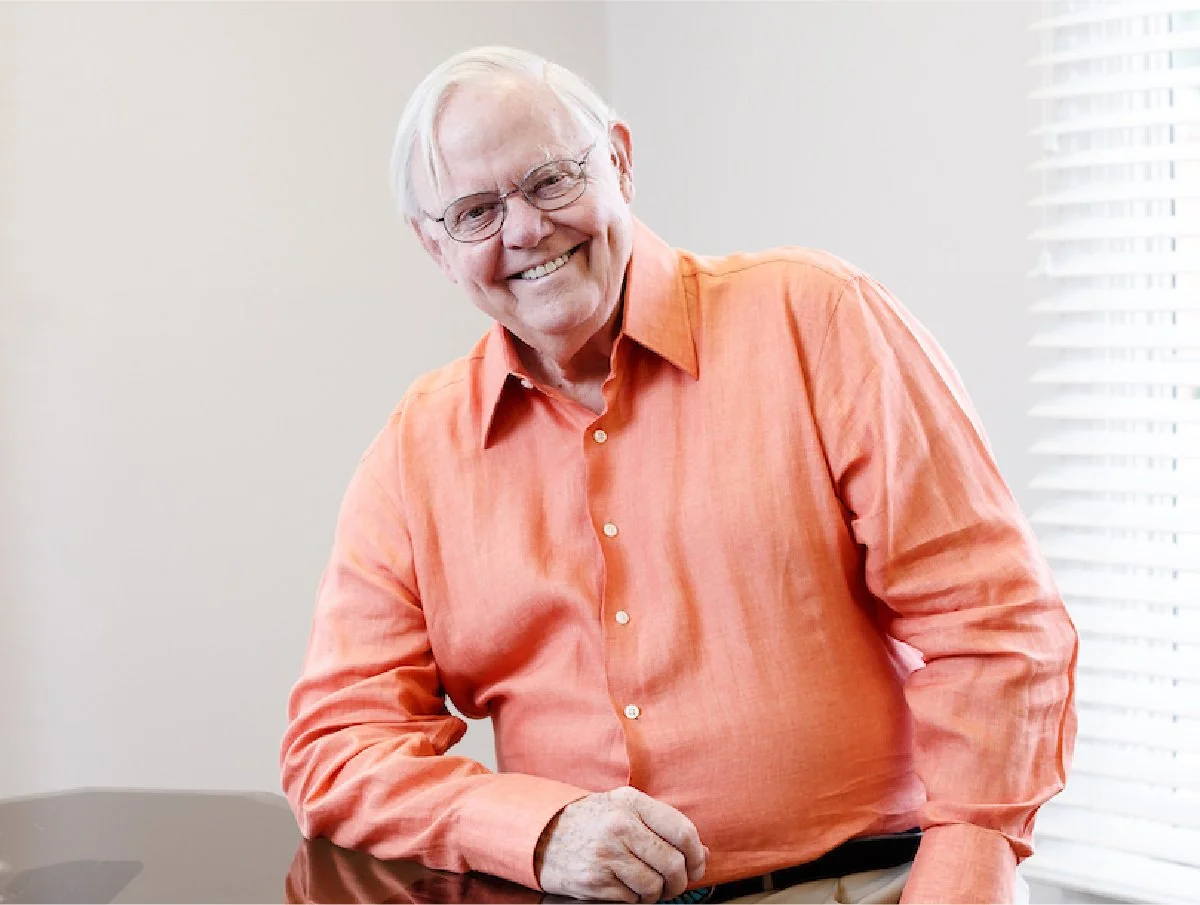 An elderly man with white hair and glasses wearing an orange button-up shirt, smiling and leaning slightly on a table.