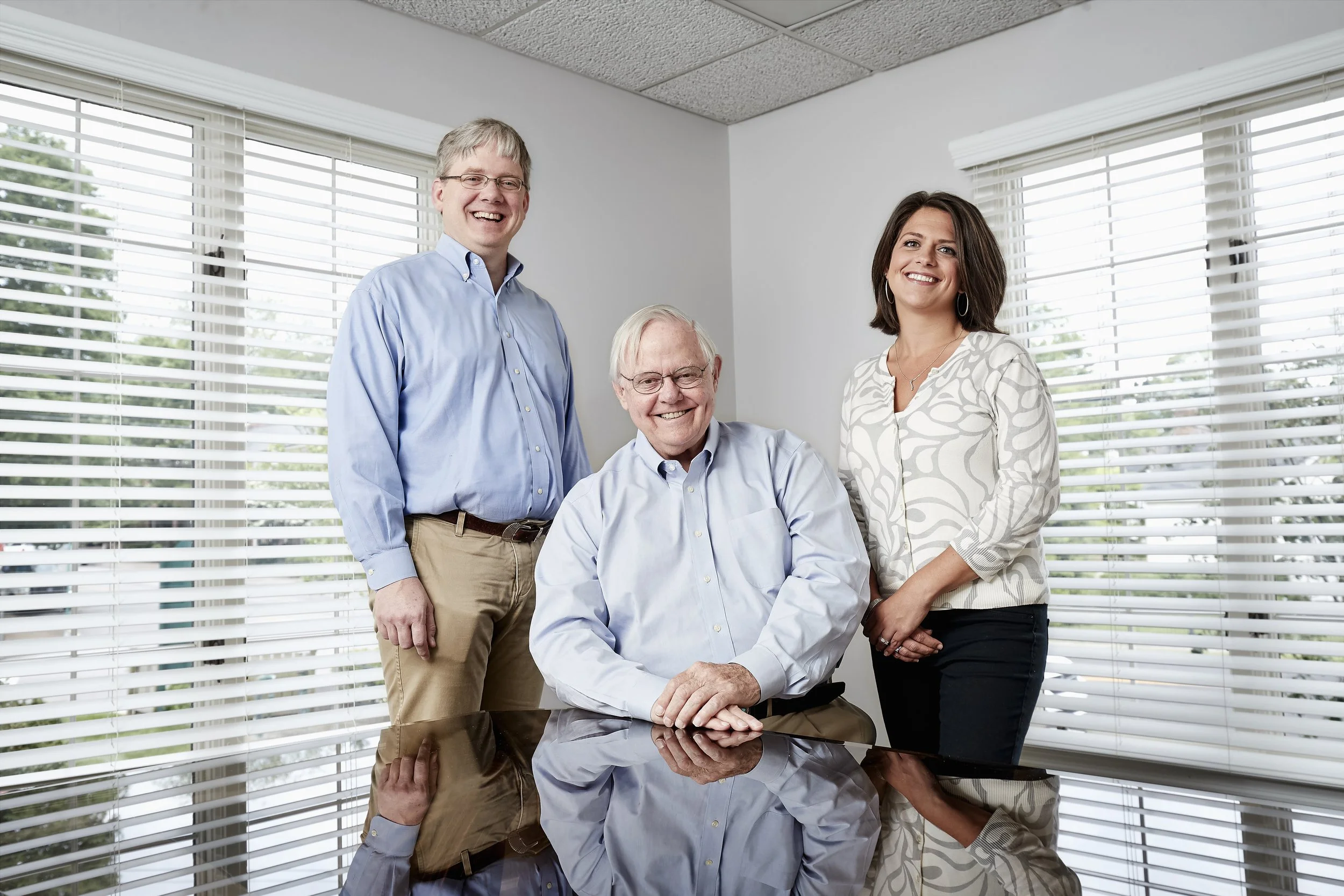 Three professionally dressed people, two men and one woman, smiling indoors with large windows and blinds in the background.
