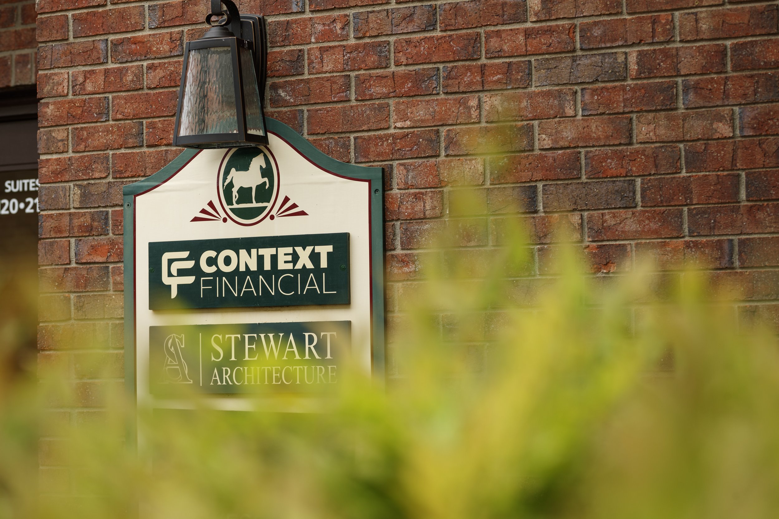 Signboard outside a building displaying logos and names of 'Context Financial' and 'Stewart Architecture' against a brick wall, with a black outdoor light fixture above it and blurred green foliage in the foreground.