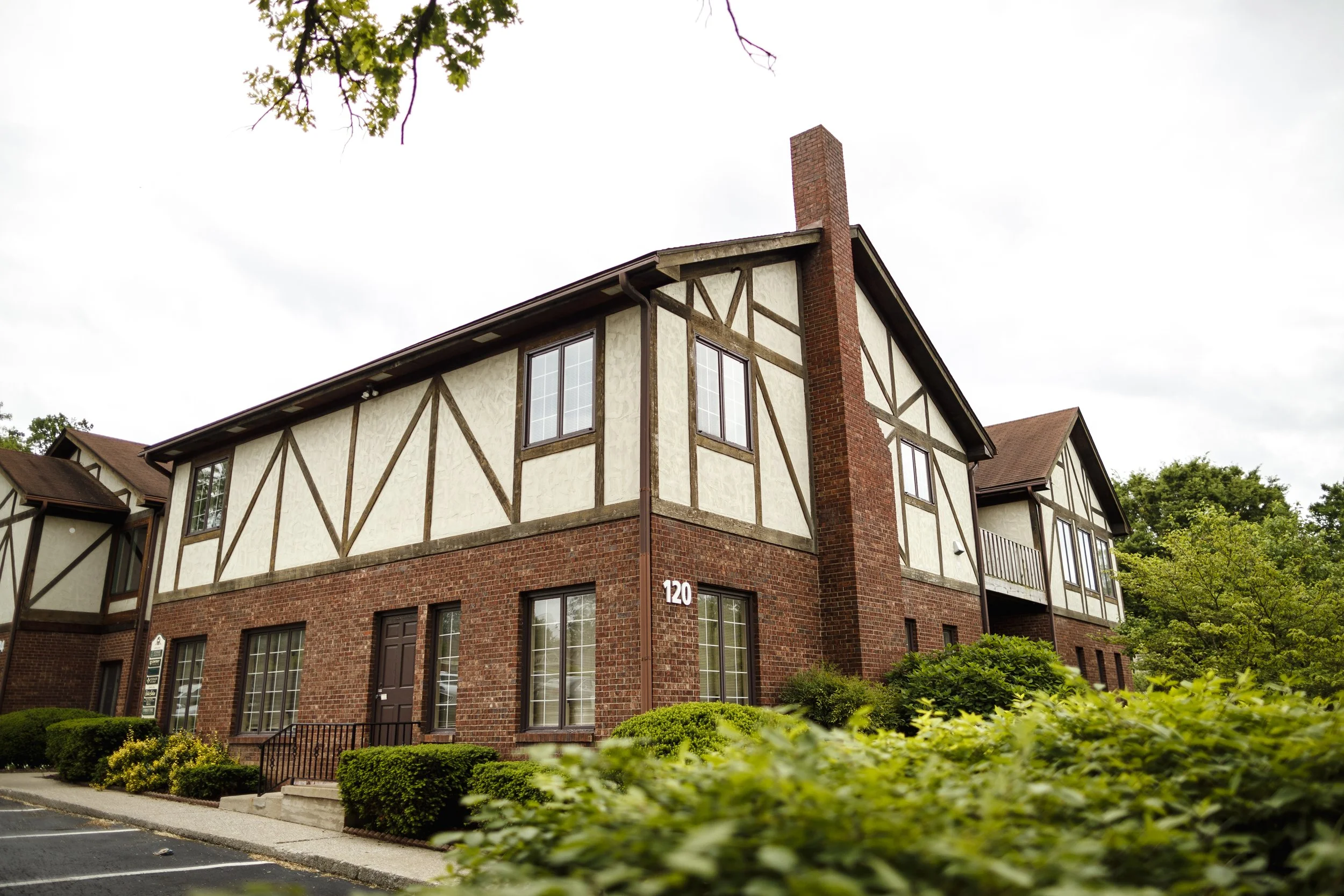 A two-story residential building with a Tudor-style exterior, brick lower section, and cream-colored upper section with dark wood trim. There are multiple windows, a small front porch with steps, and shrubbery in front. The building has a chimney and the number 120 on the wall.