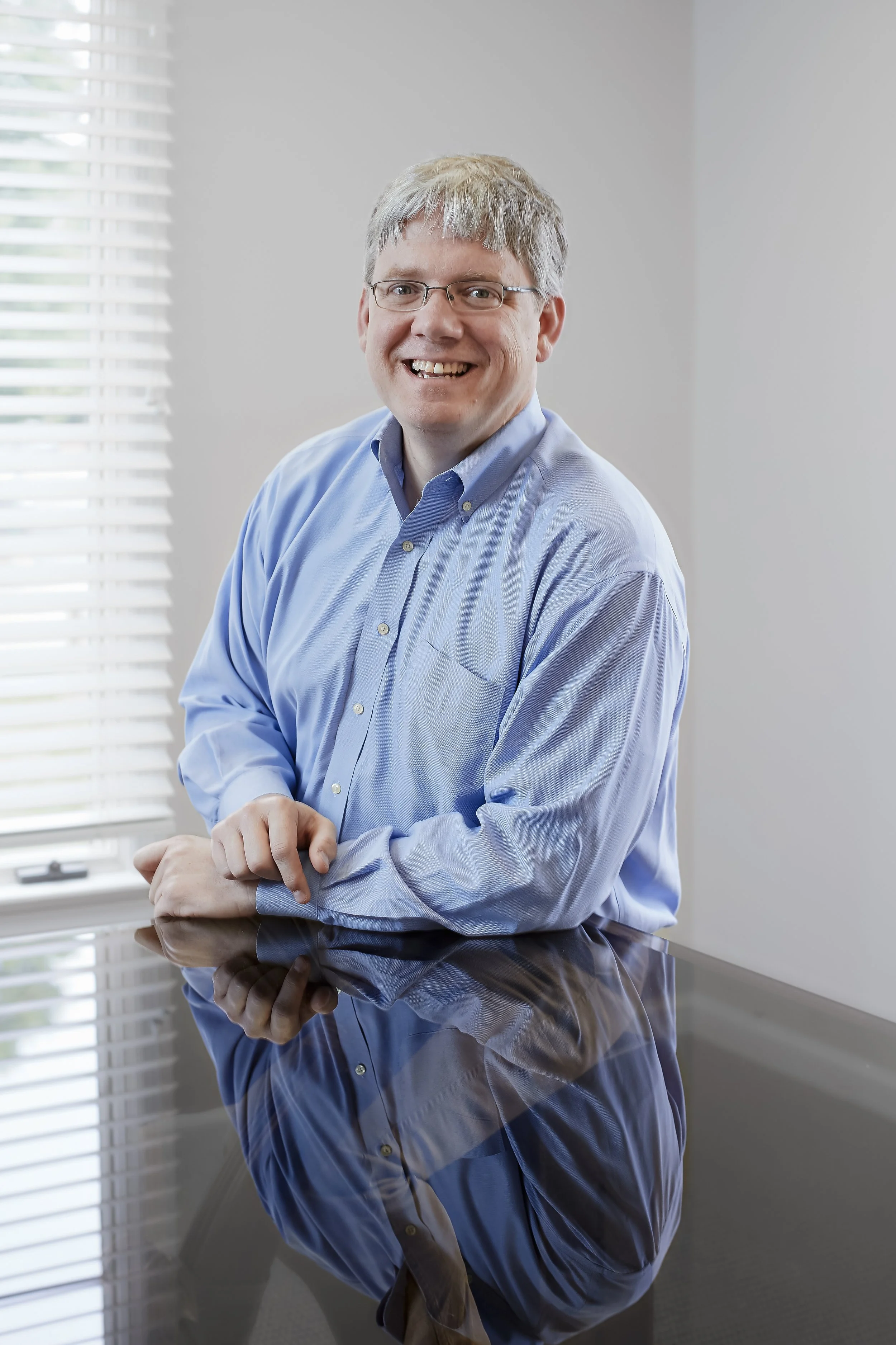 A man with glasses and gray hair, smiling, in a blue button-up shirt, sitting at a reflective black table near a window with white blinds.
