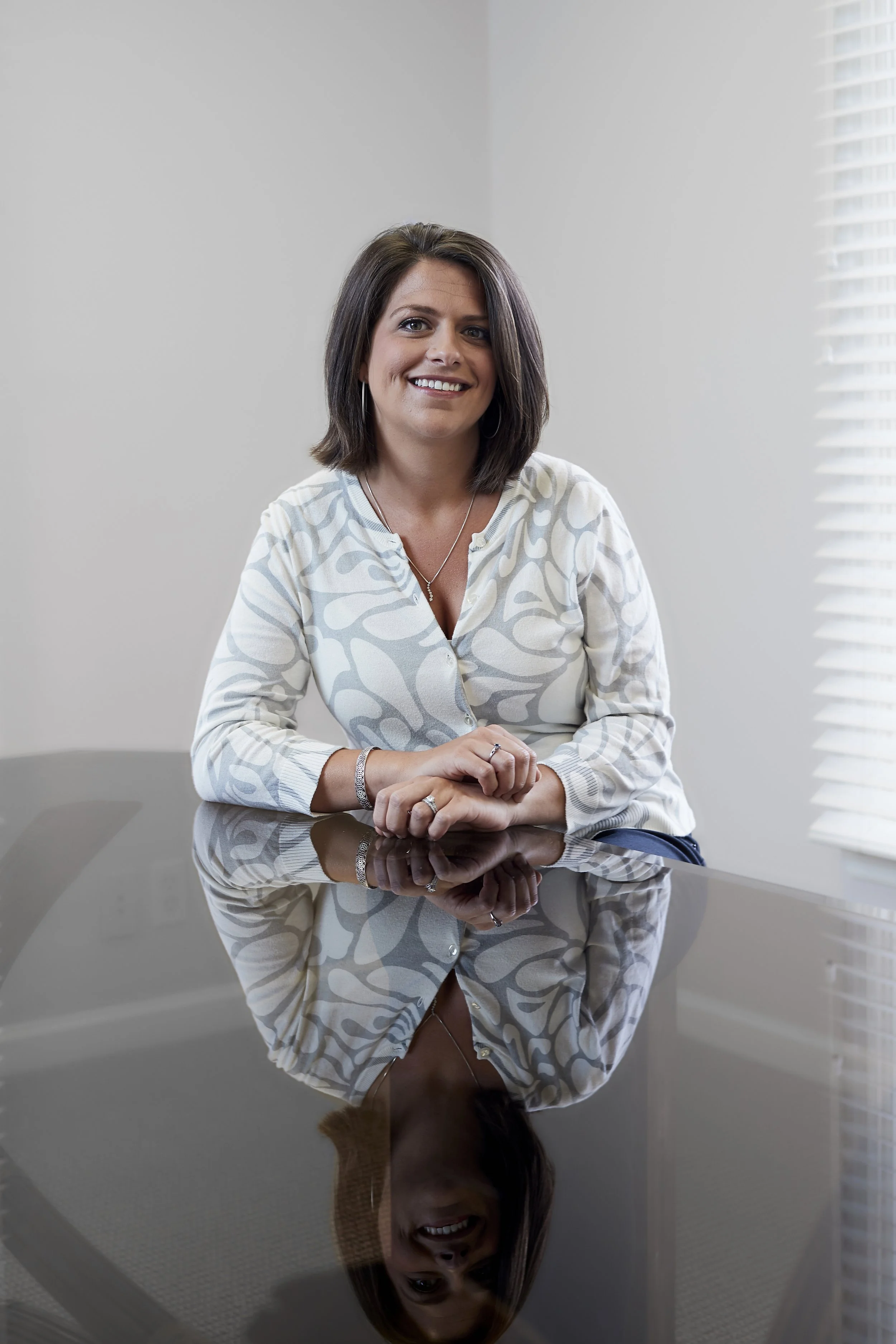 A woman sitting at a reflective black table in a bright room with white walls and blinds, smiling and looking at the camera.