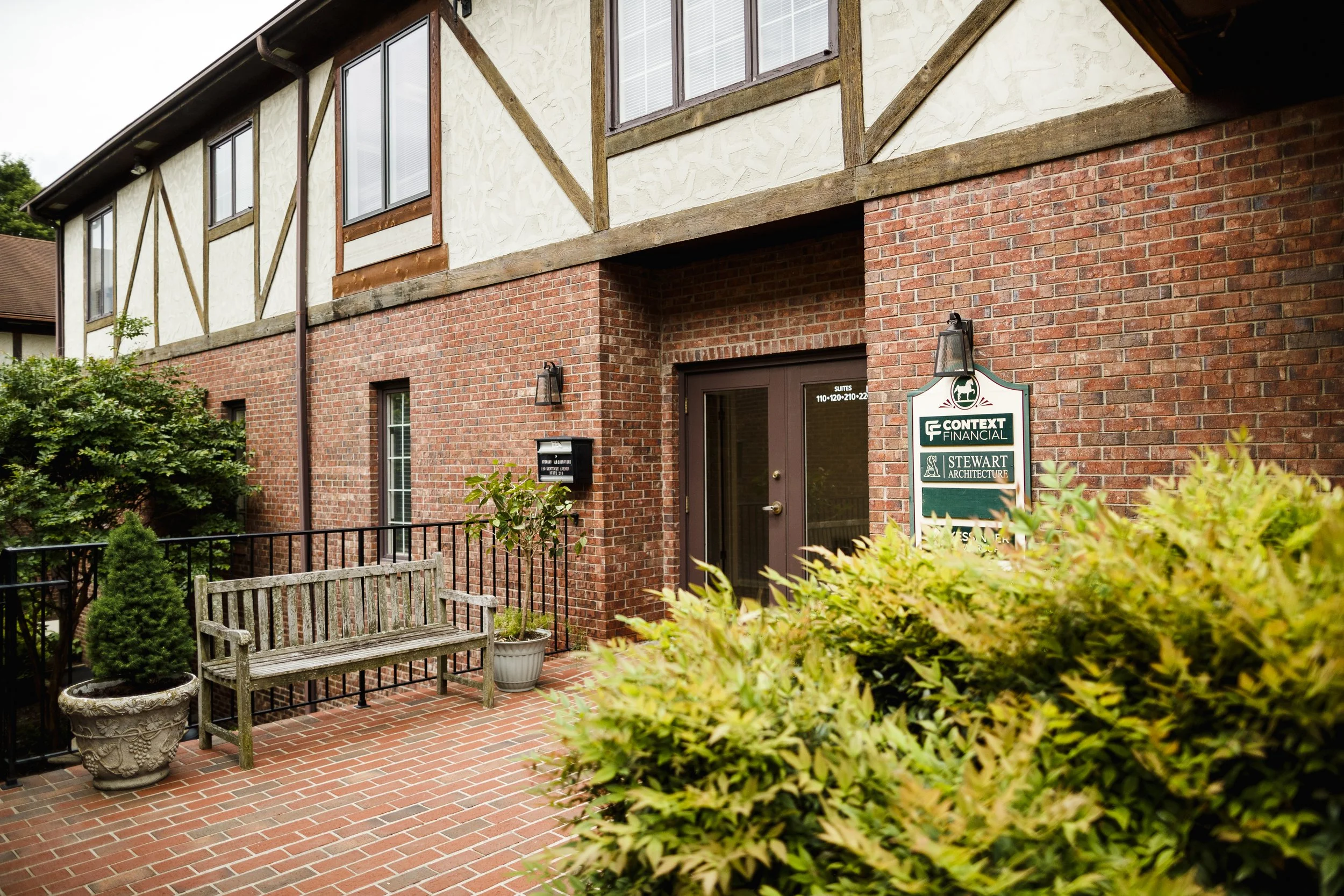 Exterior view of a brick building with a small patio area featuring a wooden bench, potted plants, and a sign listing several businesses.
