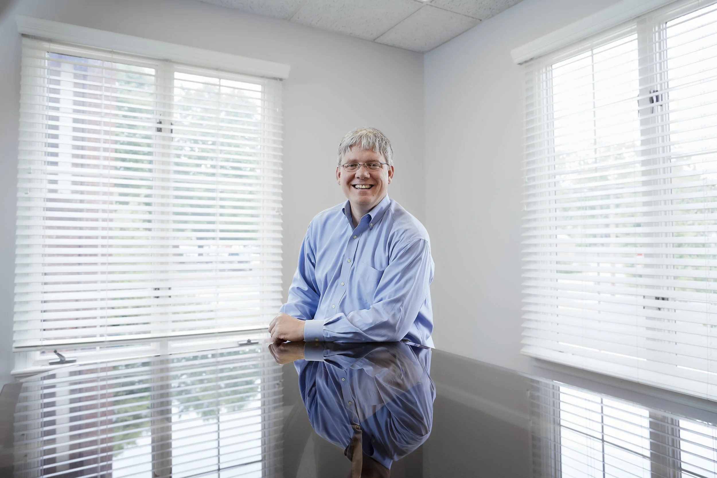 A smiling man with gray hair and glasses, wearing a light blue dress shirt, sitting at a reflective black table in a room with large windows and white blinds.
