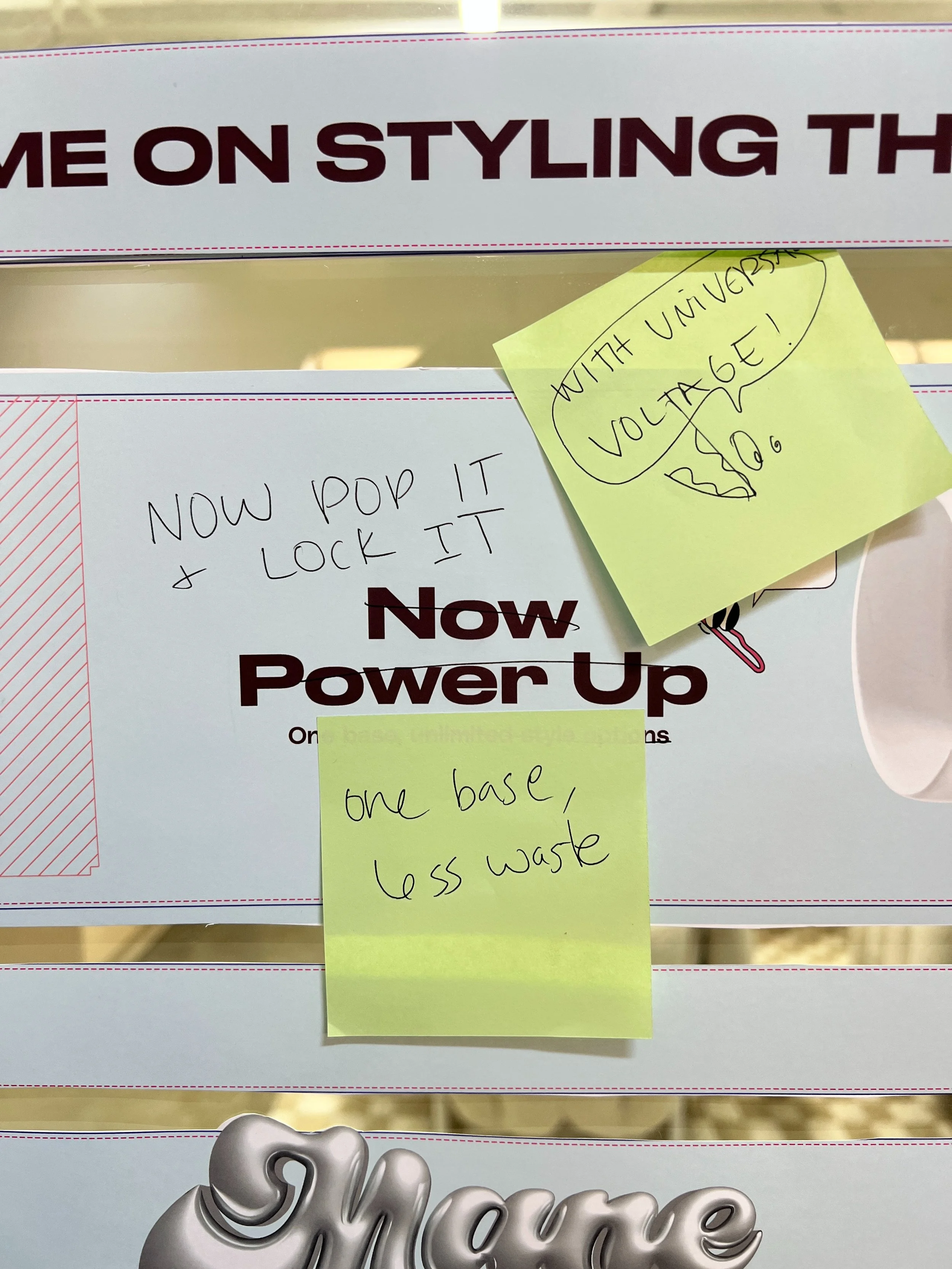Close-up of a sign with partially visible text and yellow sticky notes attached. The visible sign text reads 'ME ON STYLING THE' and 'Now Power Up'. One sticky note says 'WITH UNIVERSAL VOLTAGE!' and another says 'one base, less waste'.