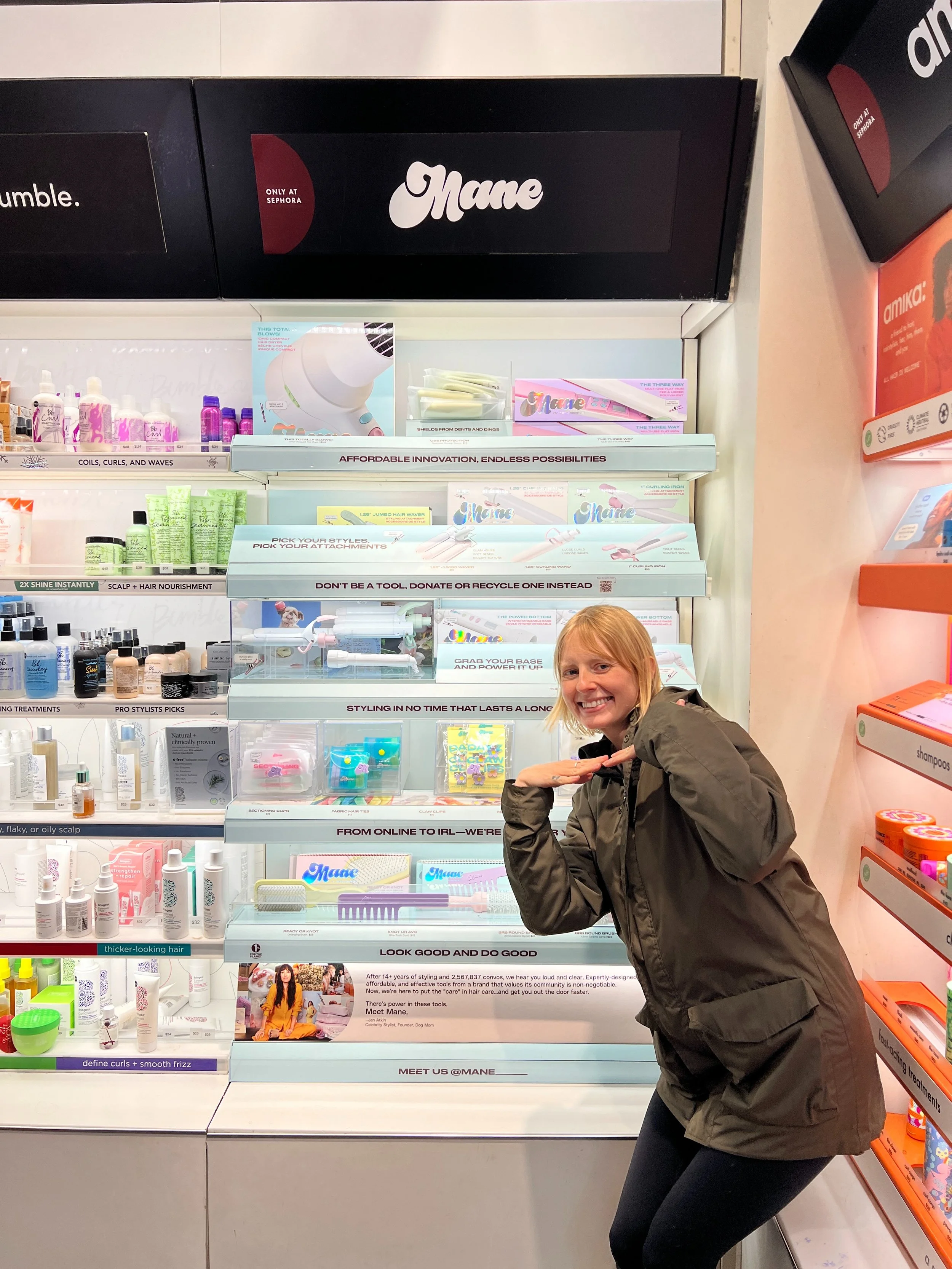 A smiling woman with blonde hair and a brown jacket crouches in front of shelves at a beauty store, holding her hands together in a pose. The shelves contain hair care products, styling tools, and accessories, with various packaging and labels visible.
