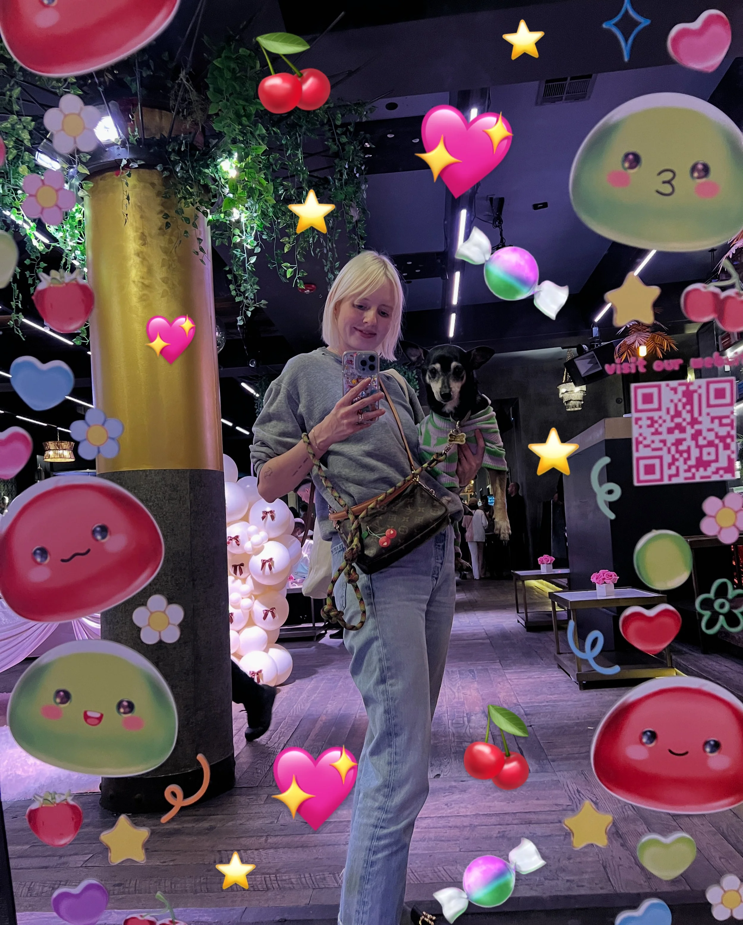 A woman with blonde hair taking a selfie with a small dog in her arms, standing in a decorated indoor space with hanging lights, colorful balloons, and various heart, star, and flower-shaped stickers overlayed on the photo.