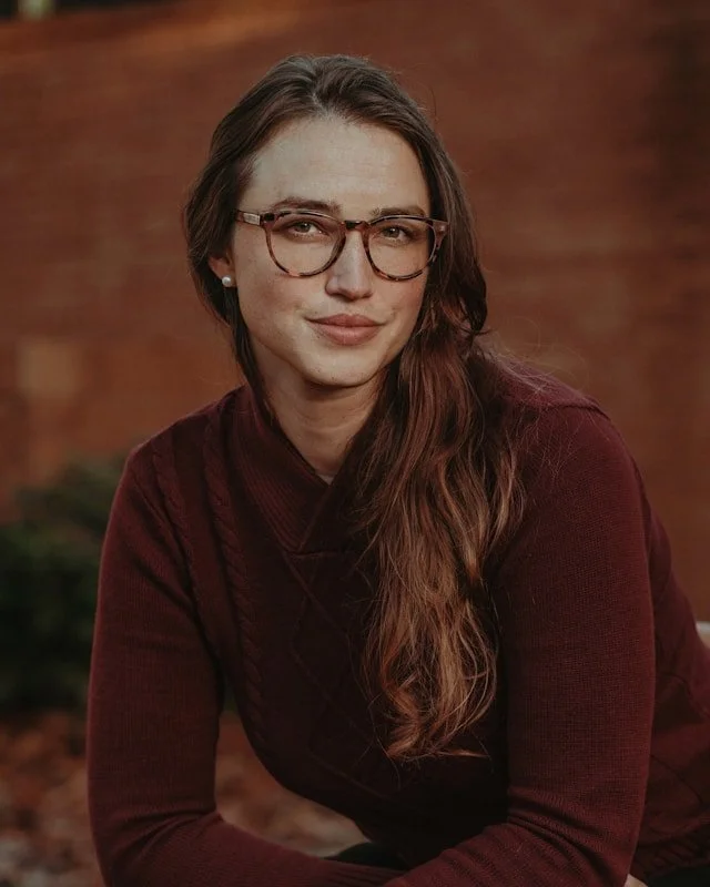 A young woman with long brown hair, wearing glasses and a dark red sweater, sitting indoors, smiling gently at the camera.