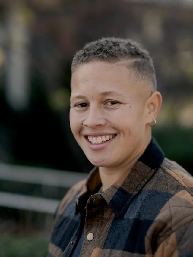 A person with short curly hair and hoop earrings smiling outdoors in front of a blurred natural background.