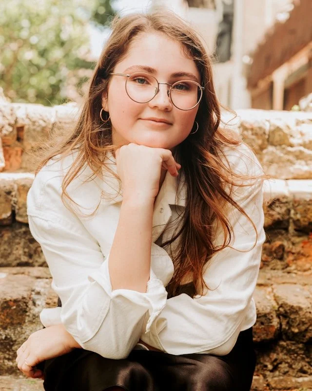 A young woman with long brown hair, glasses, and hoop earrings sitting on outdoor stone steps, resting her chin on her hand, smiling softly at the camera.