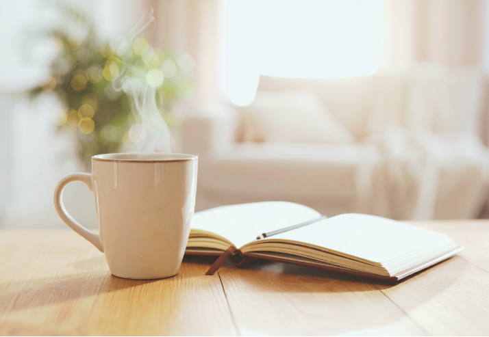 A steaming cup of coffee or tea alongside an open notebook on a wooden table with a bright living room in the background.
