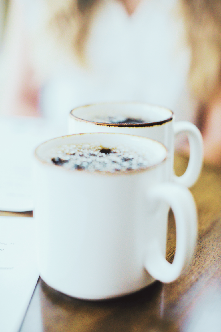 Two white mugs filled with black coffee on a wooden table, with a person wearing a white shirt blurred in the background.