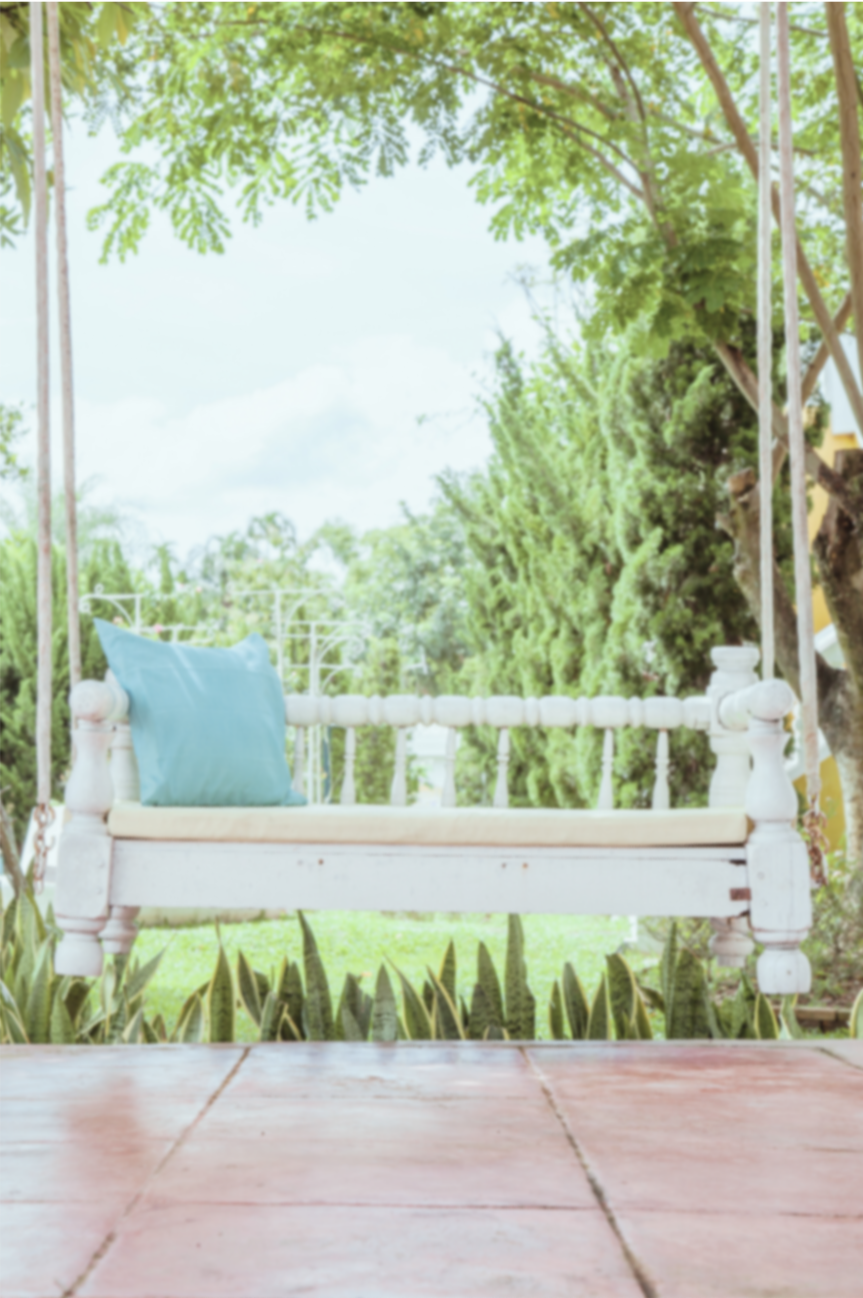 A white wooden porch swing starting with a light blue pillow hanging on one side, surrounded by lush green trees and plants outdoors during daytime.