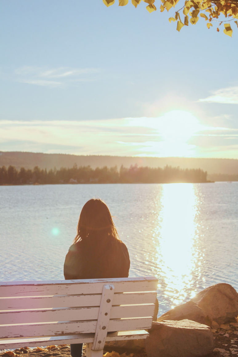 A woman with long hair sitting on a white bench by a lake, watching the sunset with a clear sky above and a tree line in the distance.