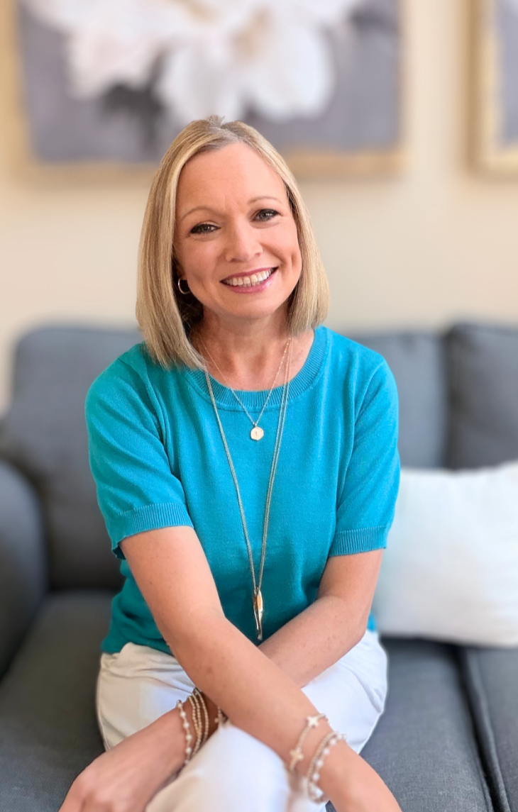 A smiling woman with shoulder-length blonde hair, wearing a bright blue short-sleeve top, white pants, and layered jewelry, sitting on a gray couch in a cozy indoor setting with framed floral artwork in the background.