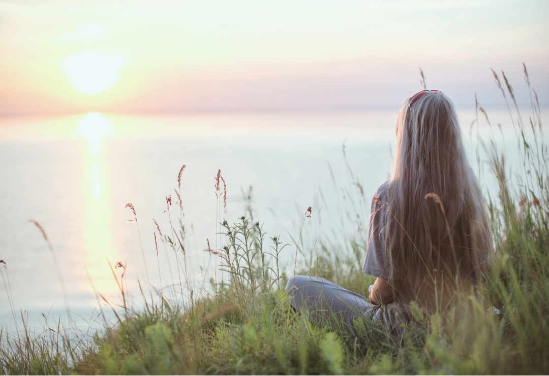 A woman sitting on grassy bank by water during sunset, facing away from camera.