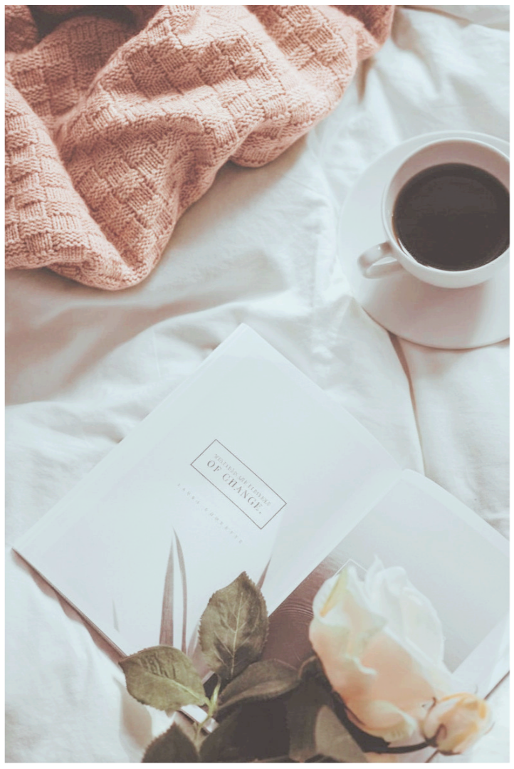 A cup of coffee on a white saucer, a pink textured blanket, an open book titled "A Handbook for Change," a white rose, and a pinkish cloth are arranged on a white bedspread.