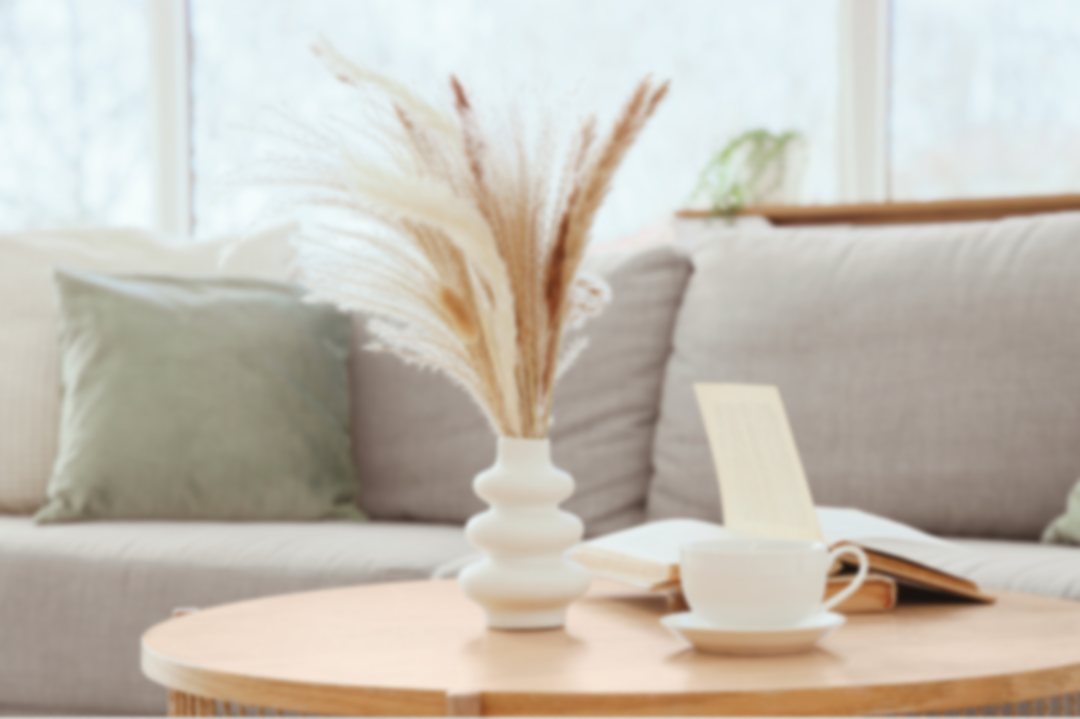 Living room with a beige sofa, green pillow, round wooden coffee table, white vase with dried grasses, open book, and a coffee cup with saucer.