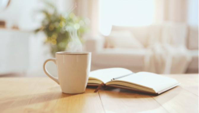 A coffee mug and an open book on a wooden table in a cozy, well-lit living room.