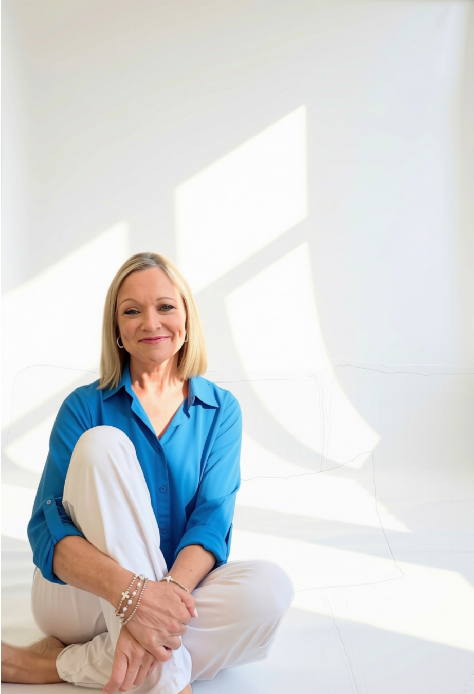 A photo of Jenni Berry sitting on the floor in a brightly lit room with sunlight streaming through window shadows, wearing a blue shirt and white pants.