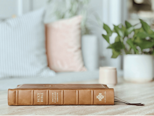 A brown leather-bound Bible resting on a wooden table in a cozy, well-lit room with pillows and plants in the background.