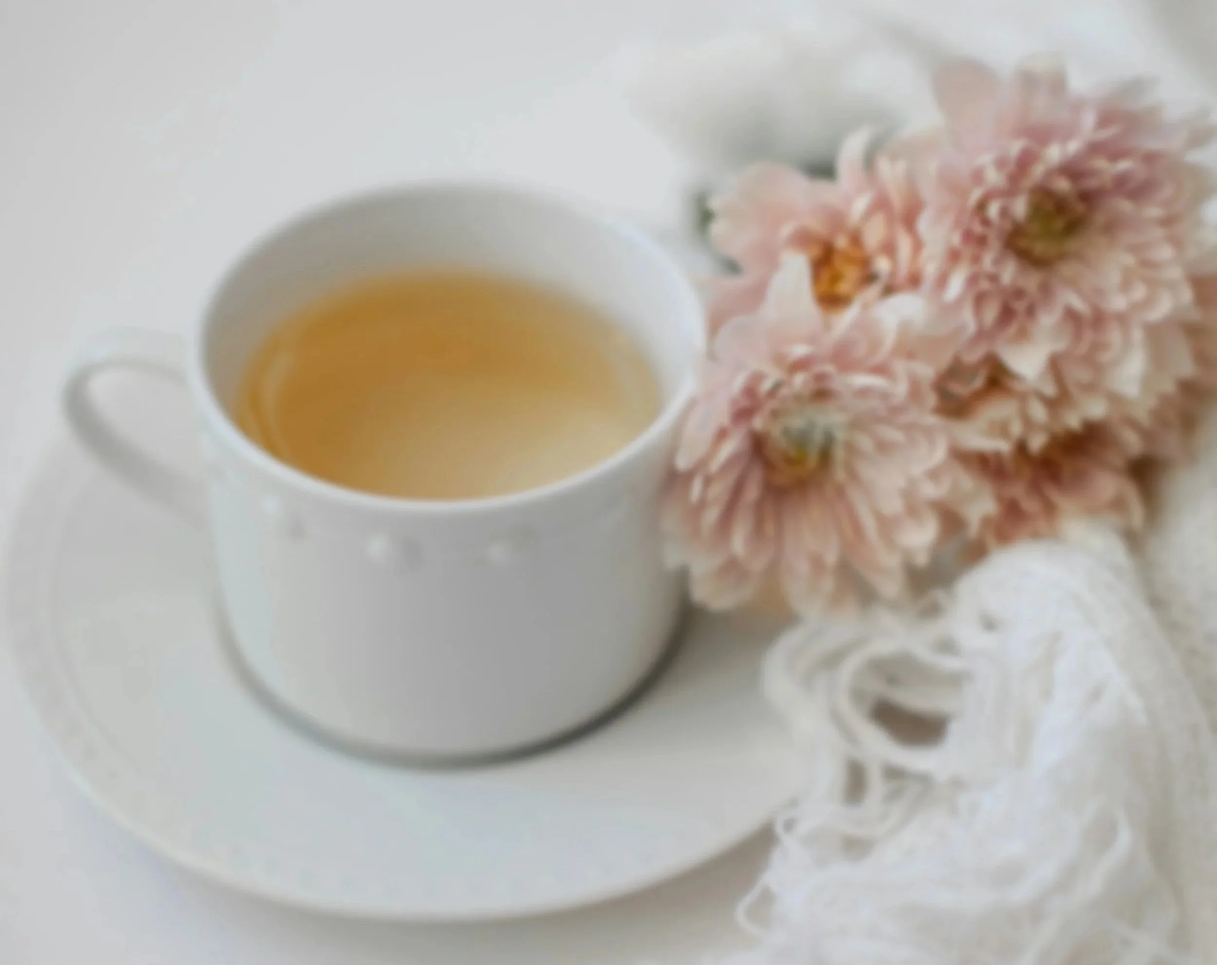Close-up of a white cup filled with tea next to a pink chrysanthemum flower on a white surface.