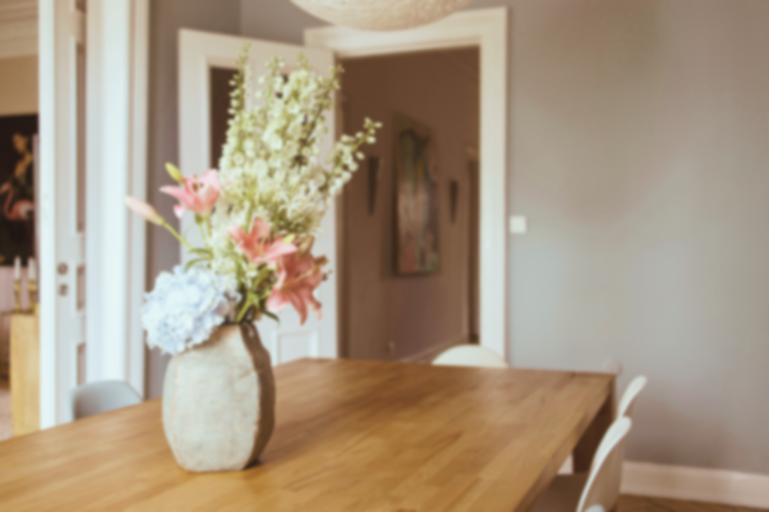 A bouquet of pink and white flowers in a rustic beige vase on a wooden dining table in a modern, cozy dining room.