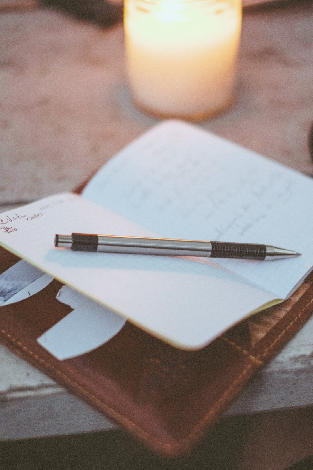 Open notebook with handwritten notes and a black pen resting on it. The notebook is on top of a leather-bound journal or book, placed on a table. In the background, there is a lit candle creating a warm glow.