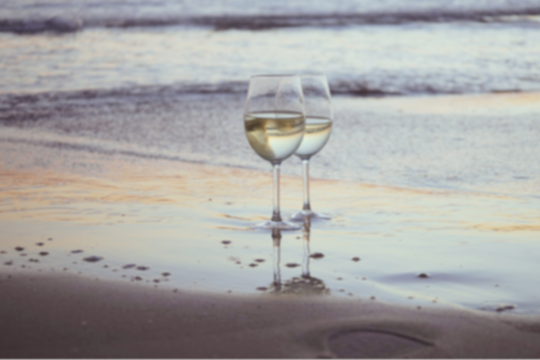 Two wine glasses filled with white wine placed on the beach at the shoreline during sunset.