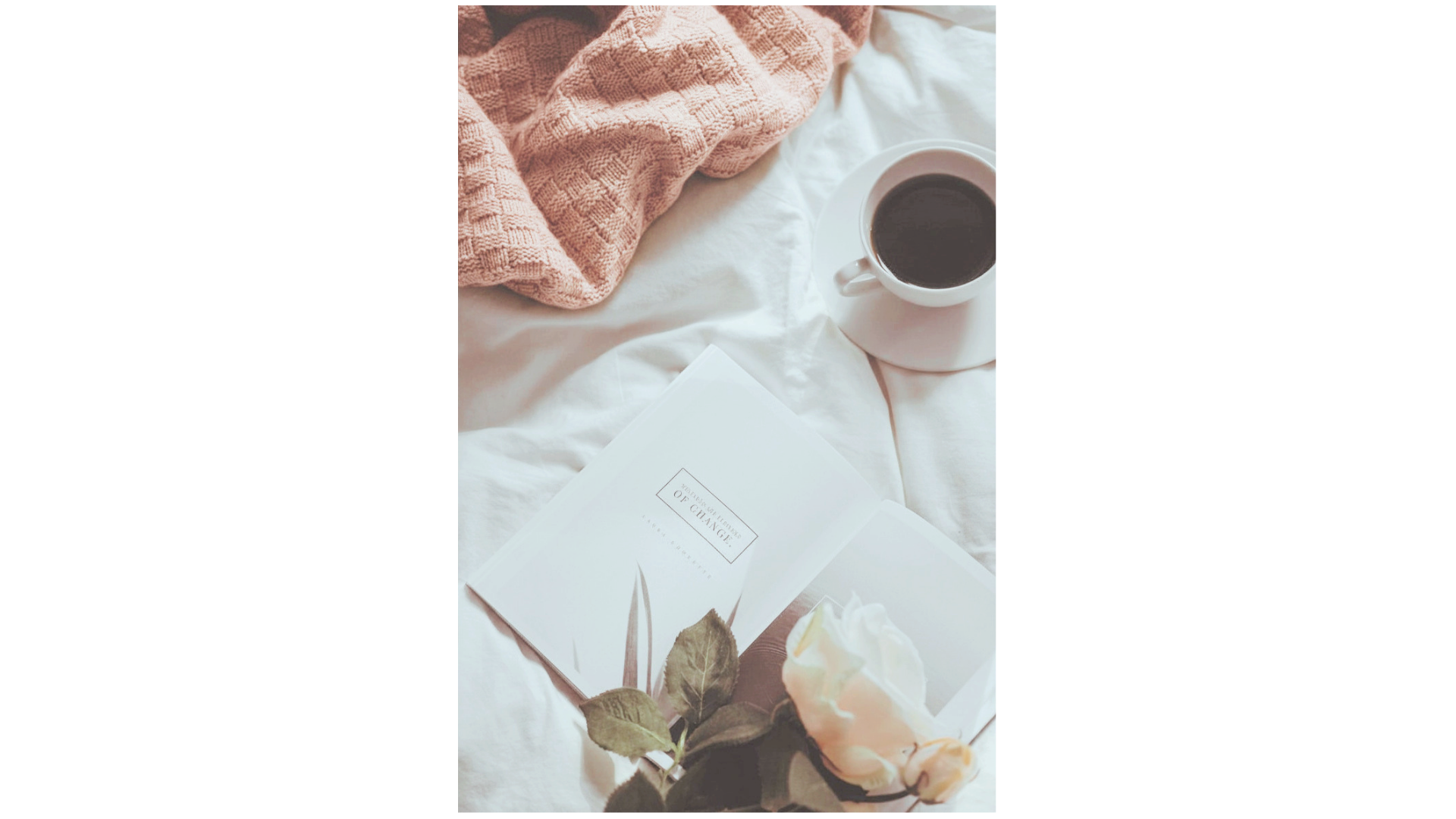 A cozy scene with a pink knitted blanket, a white cup of black coffee, an open book, a white flower, and some dried leaves on a soft white surface.