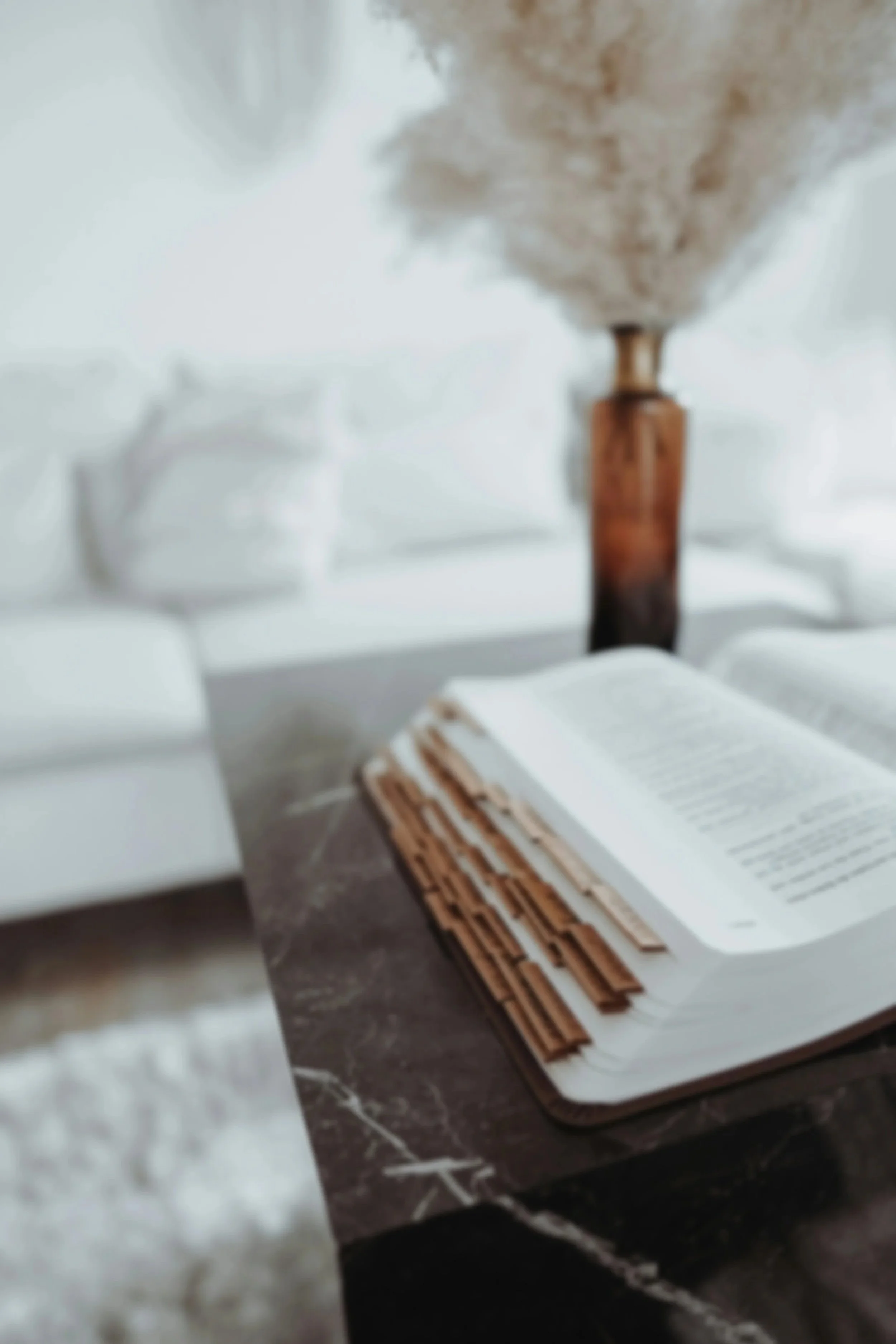 A stack of newspapers or magazines on a dark surface in front of a blurred background with a tall, ceramic vase with dried or preserved flowers or pampas grass.
