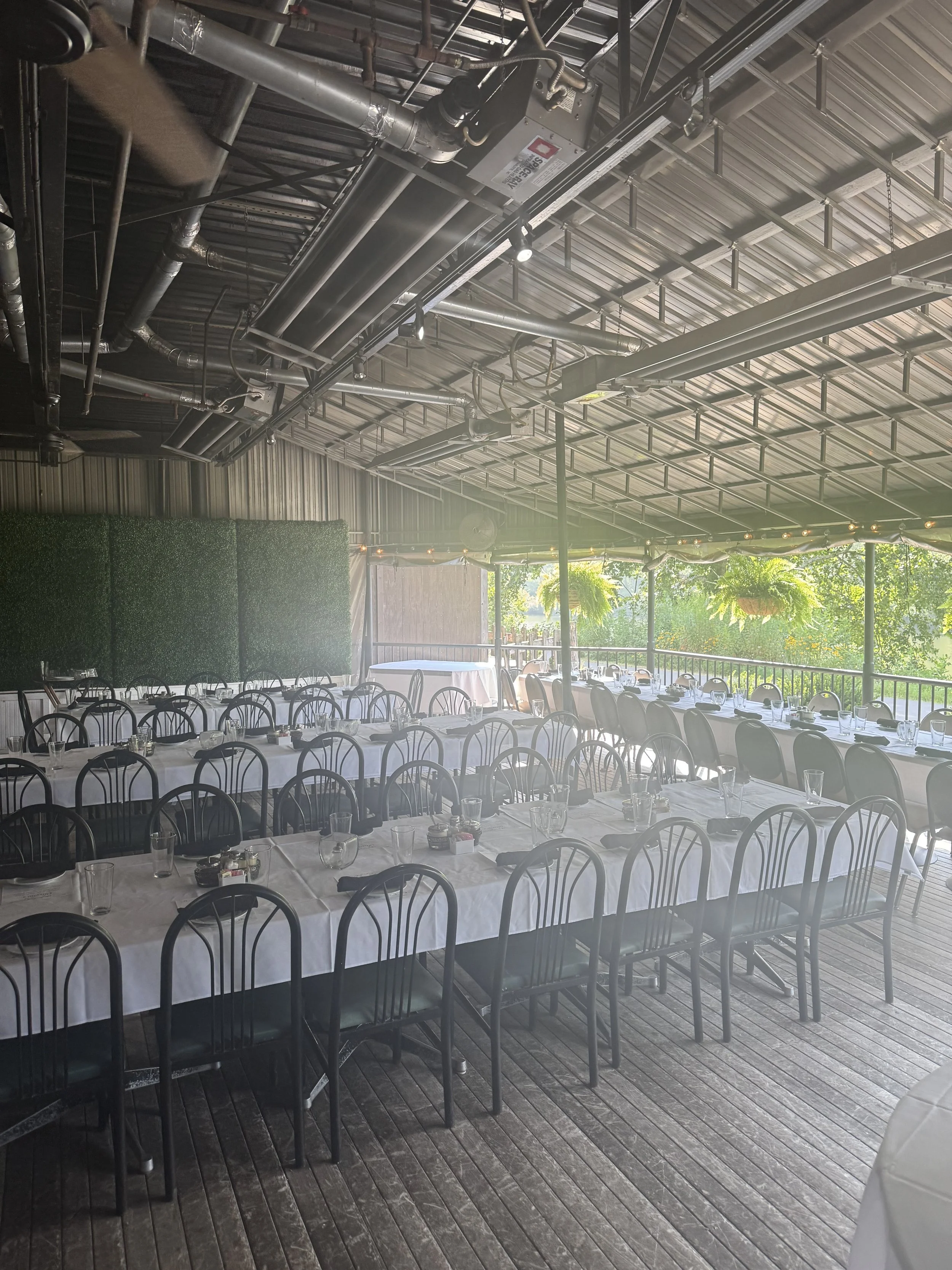 Set dining tables with white tablecloths, glasses, and utensils in an outdoor covered pavilion with string lights and greenery.