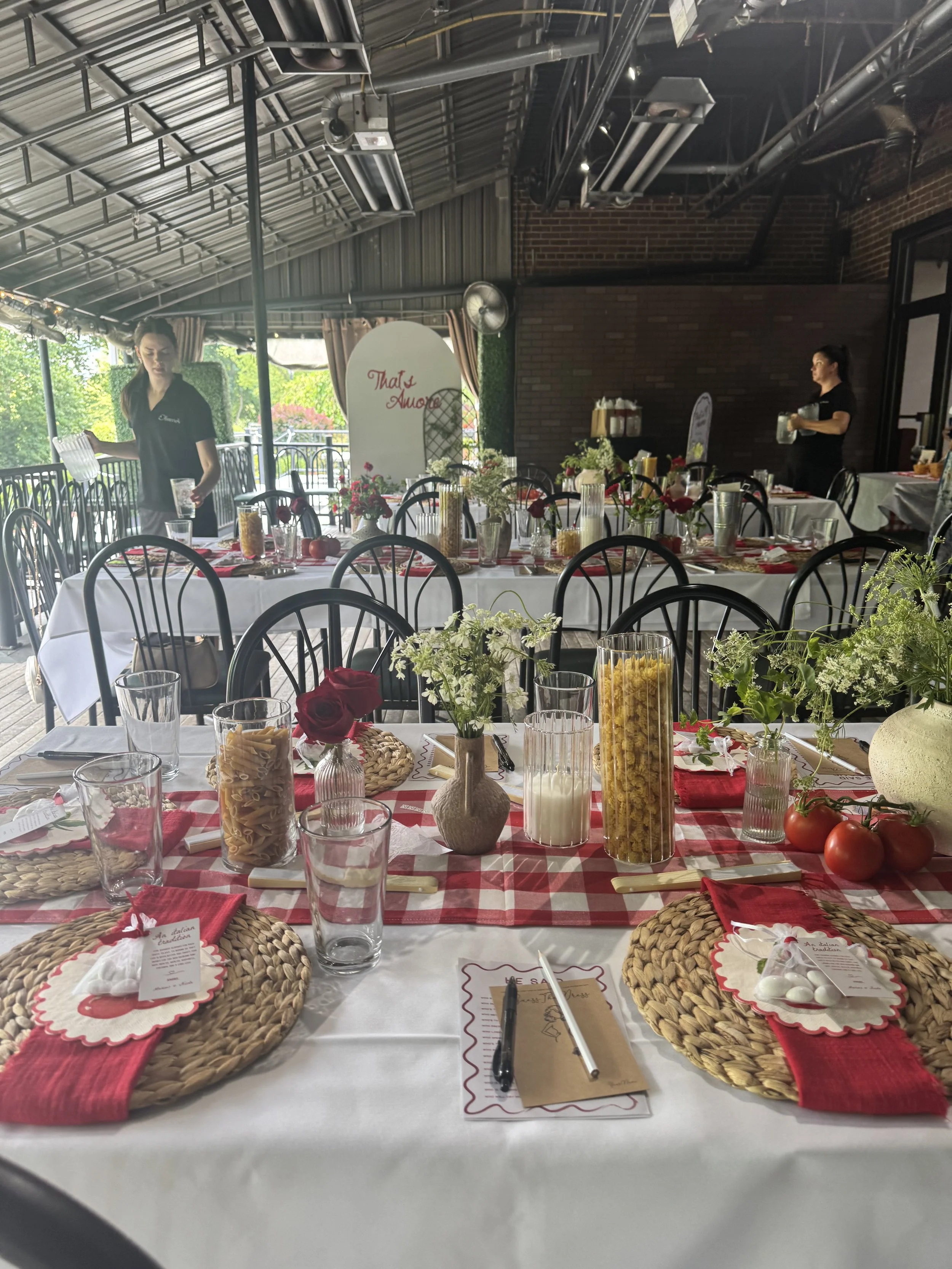 A decorated dining table set for a meal, with a red and white checkered runner, woven placemats, red napkins, clear glasses, vases with flowers, and various containers of pasta and tomatoes, in a room with a metal ceiling and two women in black shirts preparing or serving.