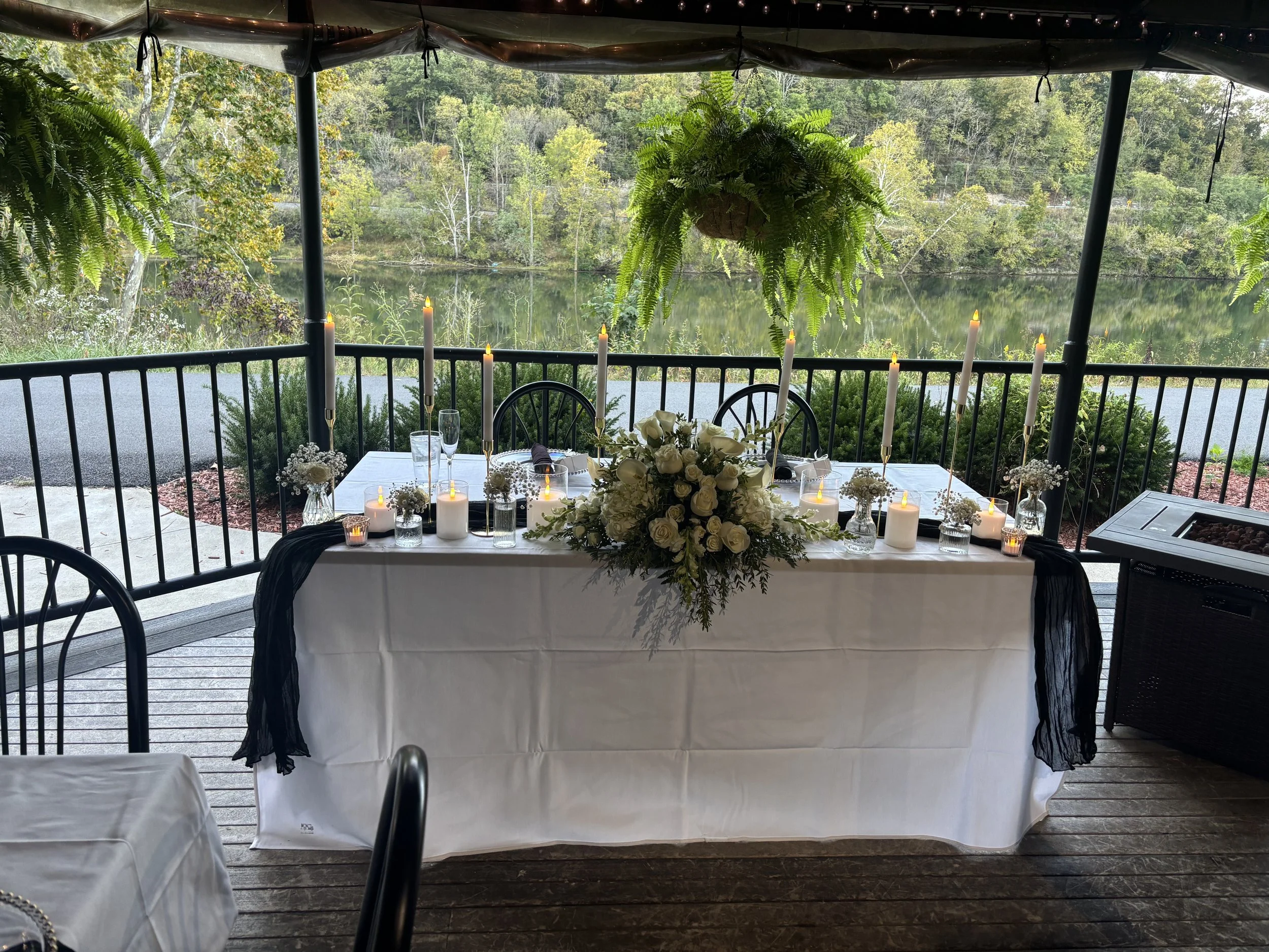 A decorated outdoor table with white tablecloth, white roses, candles, and hanging ferns, overlooking a lake and green trees.