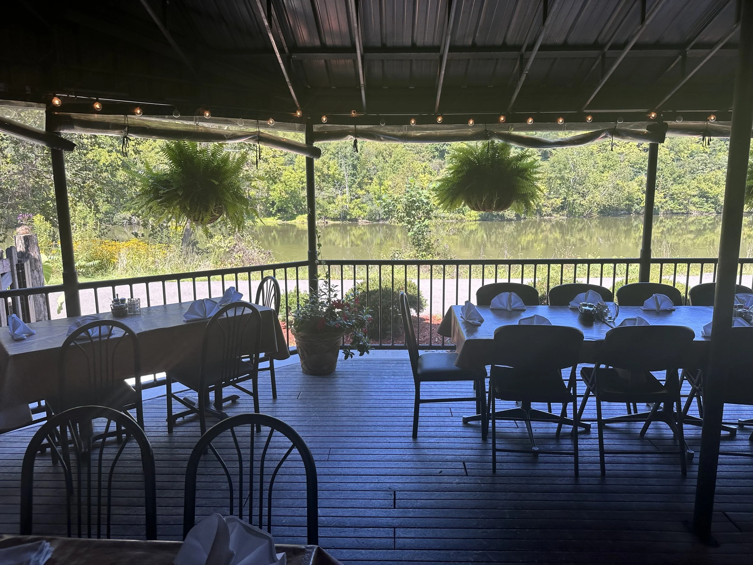 Outdoor dining area with tables set with napkins and salt and pepper shakers, overlooking a lake with green trees and grass, under a roof with hanging plants and string lights.