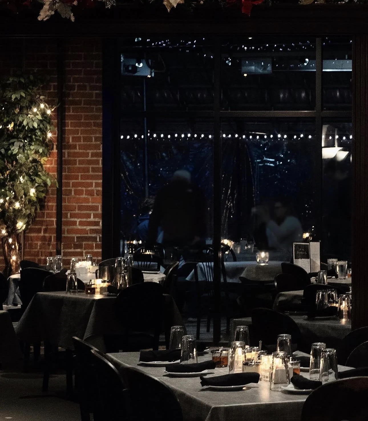 A dimly lit restaurant with tables set with black napkins and glasses, candles on the tables, a brick wall on the left, and string lights outside a window in the background.