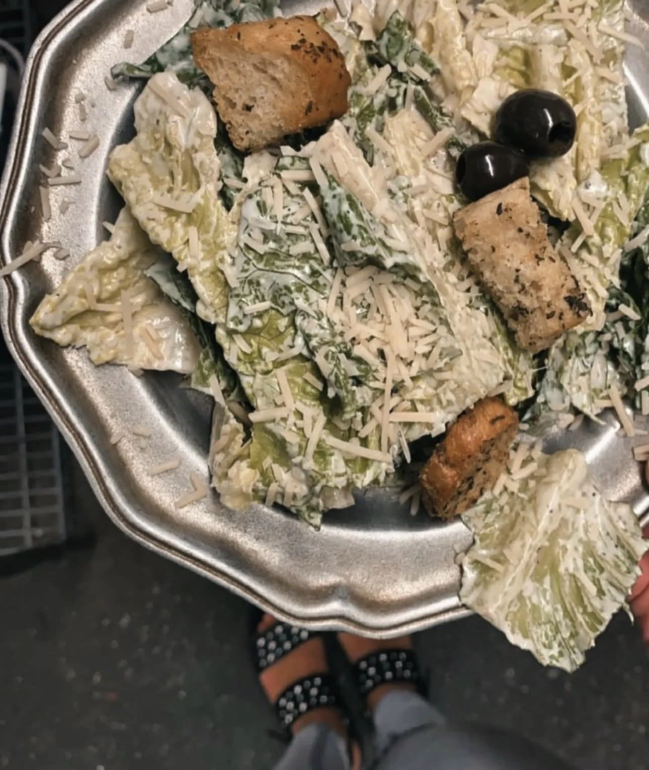 A metal tray holding a Caesar salad with romaine lettuce, grated cheese, grilled chicken pieces, black olives, and croutons.