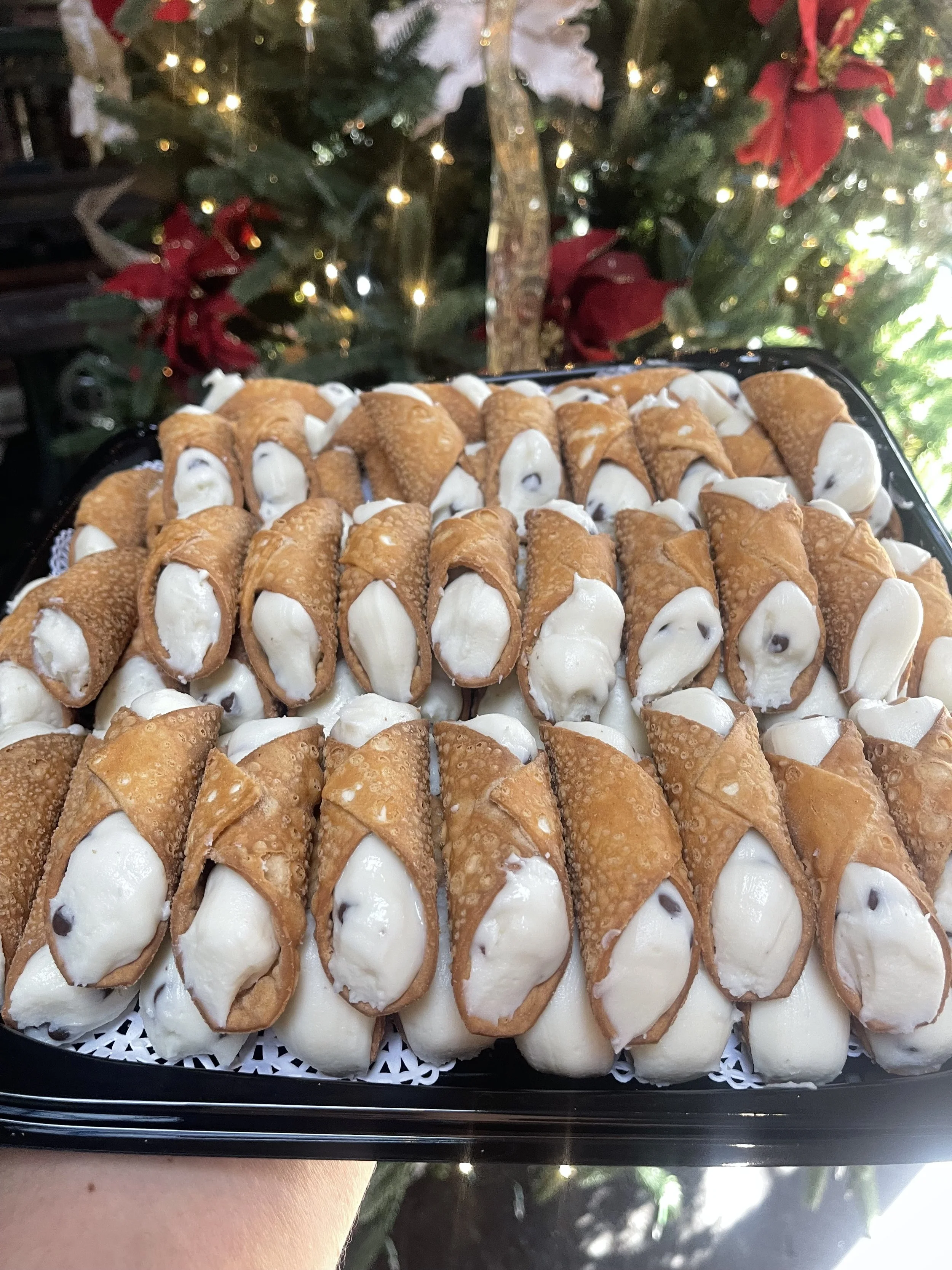 Tray of cannoli with creamy filling and chocolate chips, set against a Christmas tree with decorations and lights in the background.