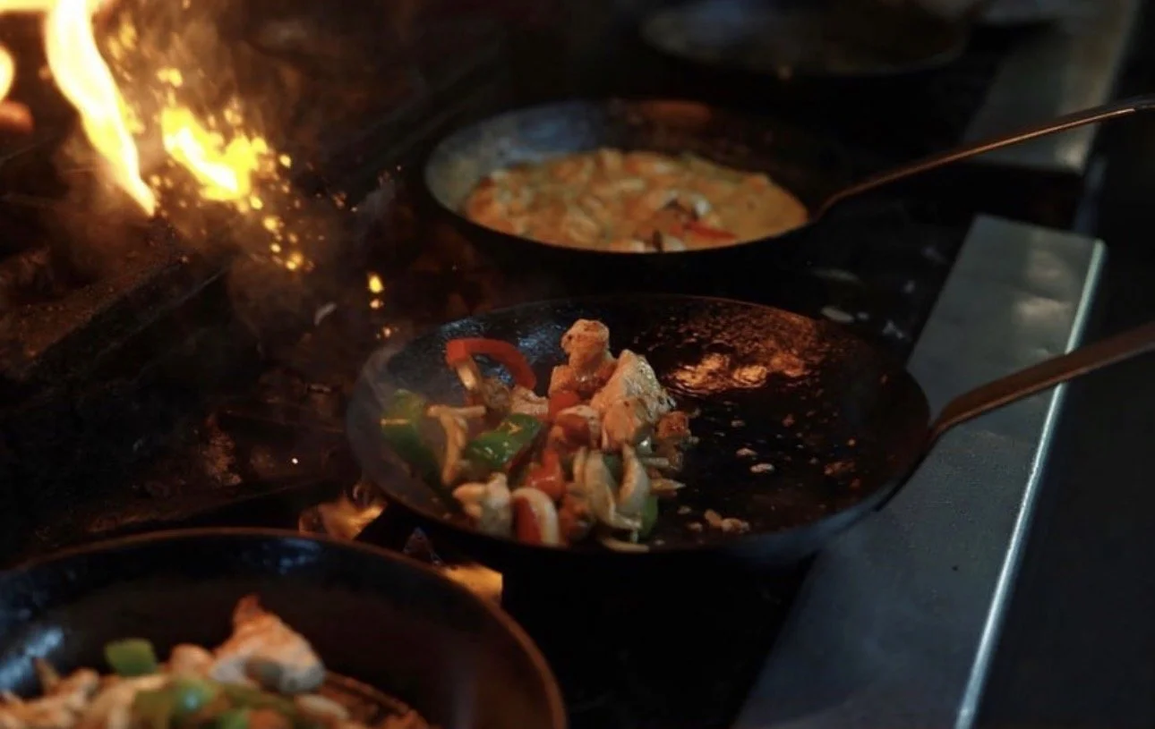 Pans with sizzling vegetables and ingredients on a stove, with flames visible at the back.