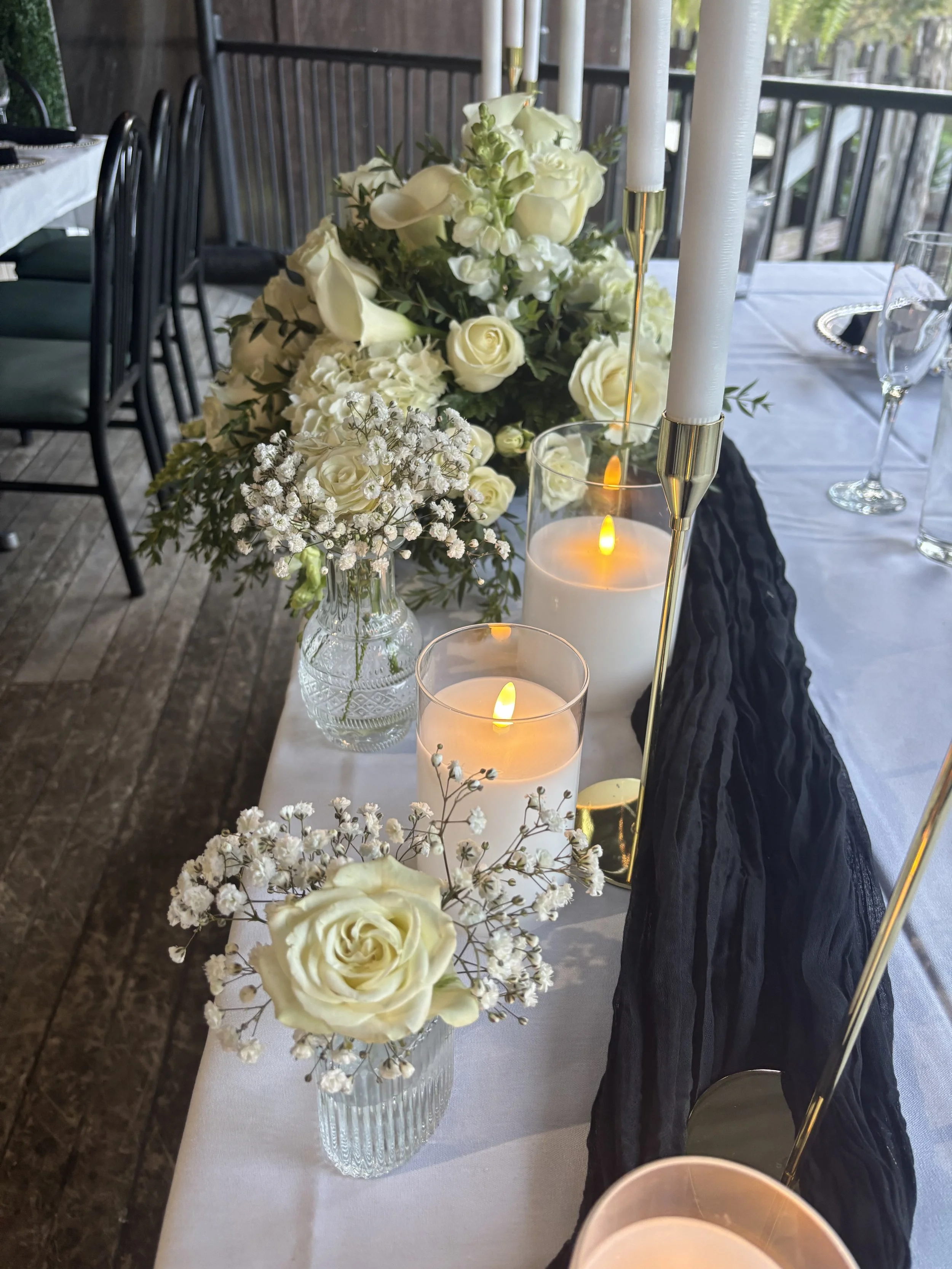 Elegant dining table with white roses, baby's breath, candles, and black and white tablecloths in a decorated setting.