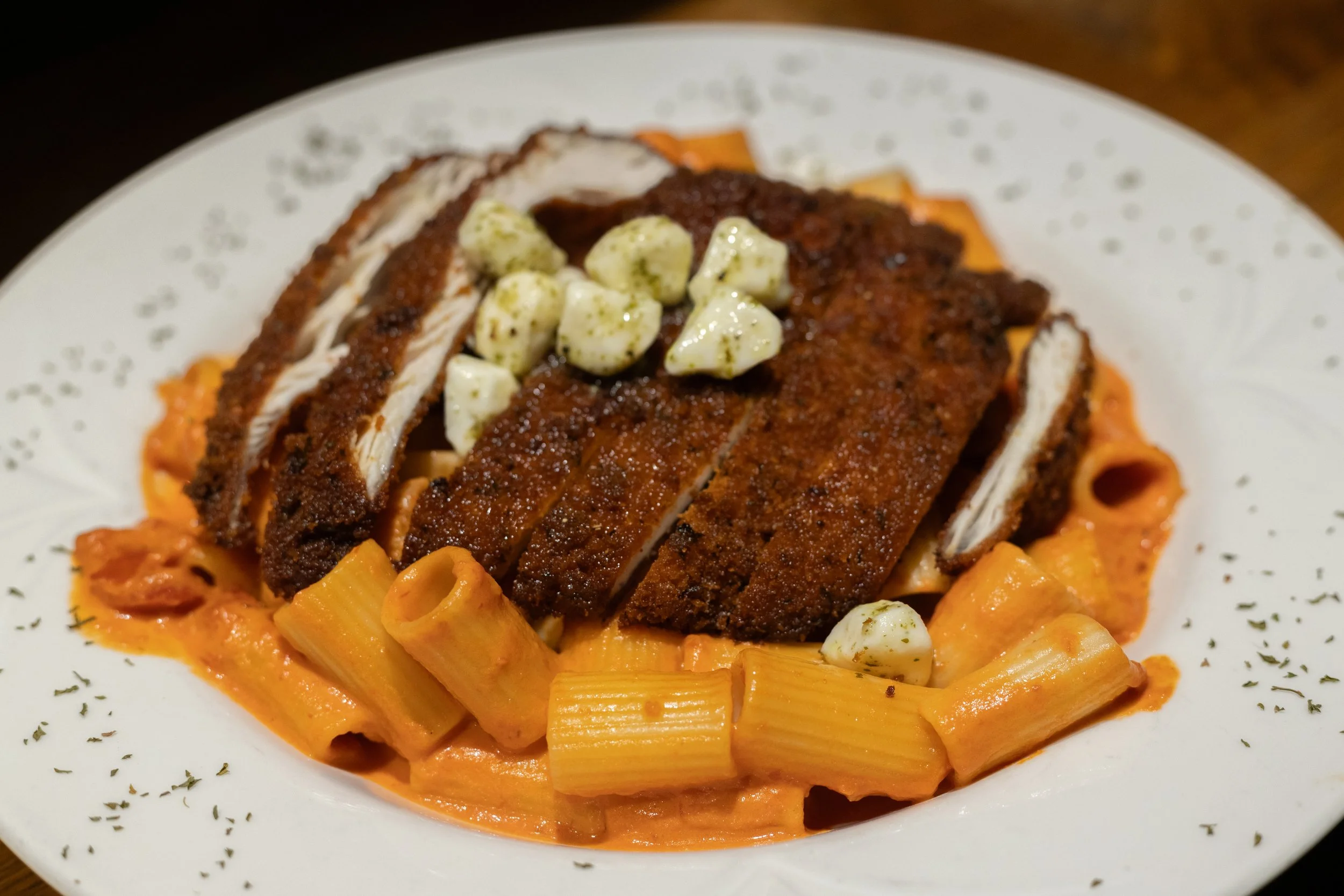 Plate of pasta with tomato sauce, topped with sliced breaded fried chicken, garnished with chunks of cheese and herbs.