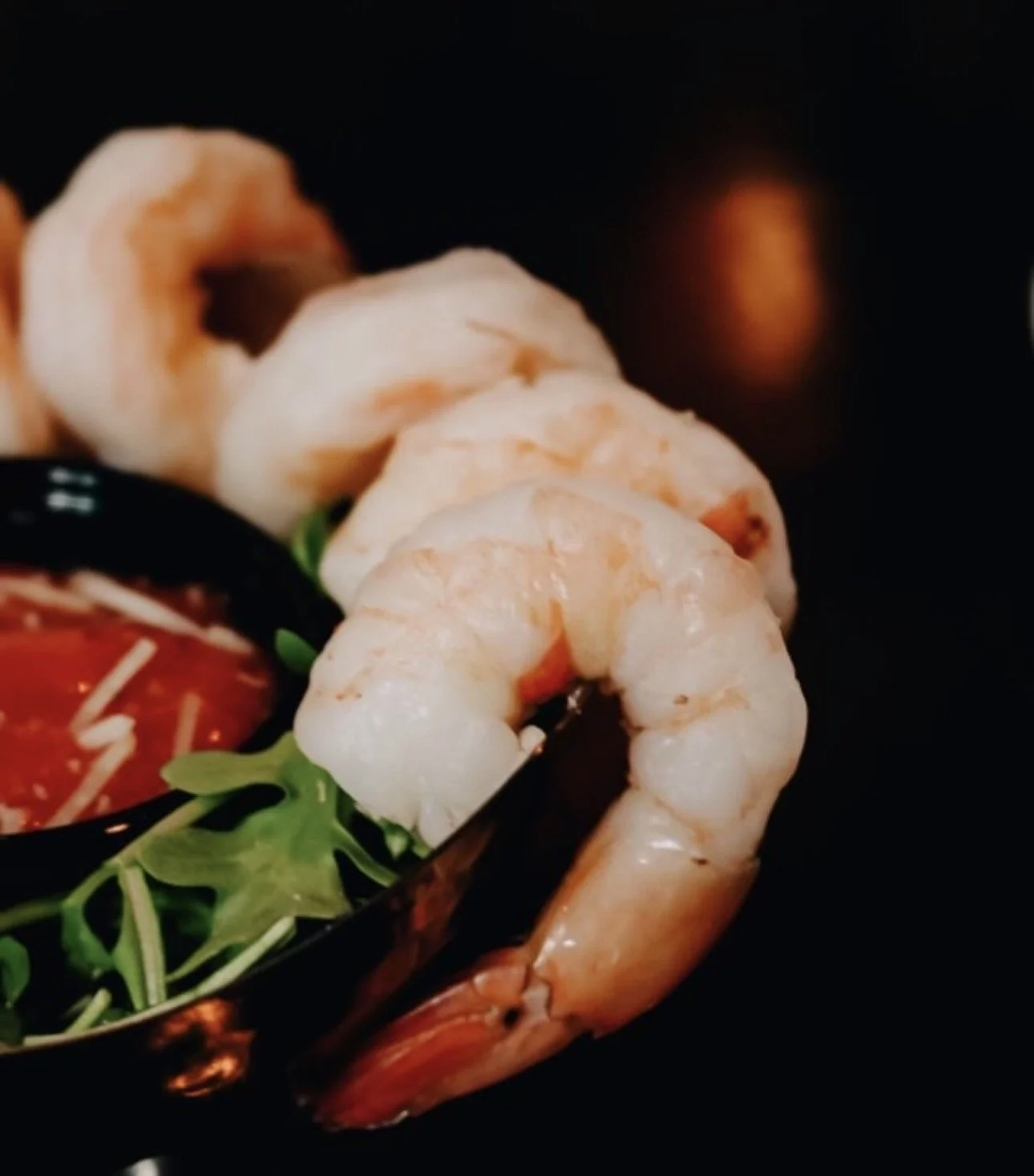 Close-up of cooked shrimp arranged with greens and tomato sauce in a black dish.