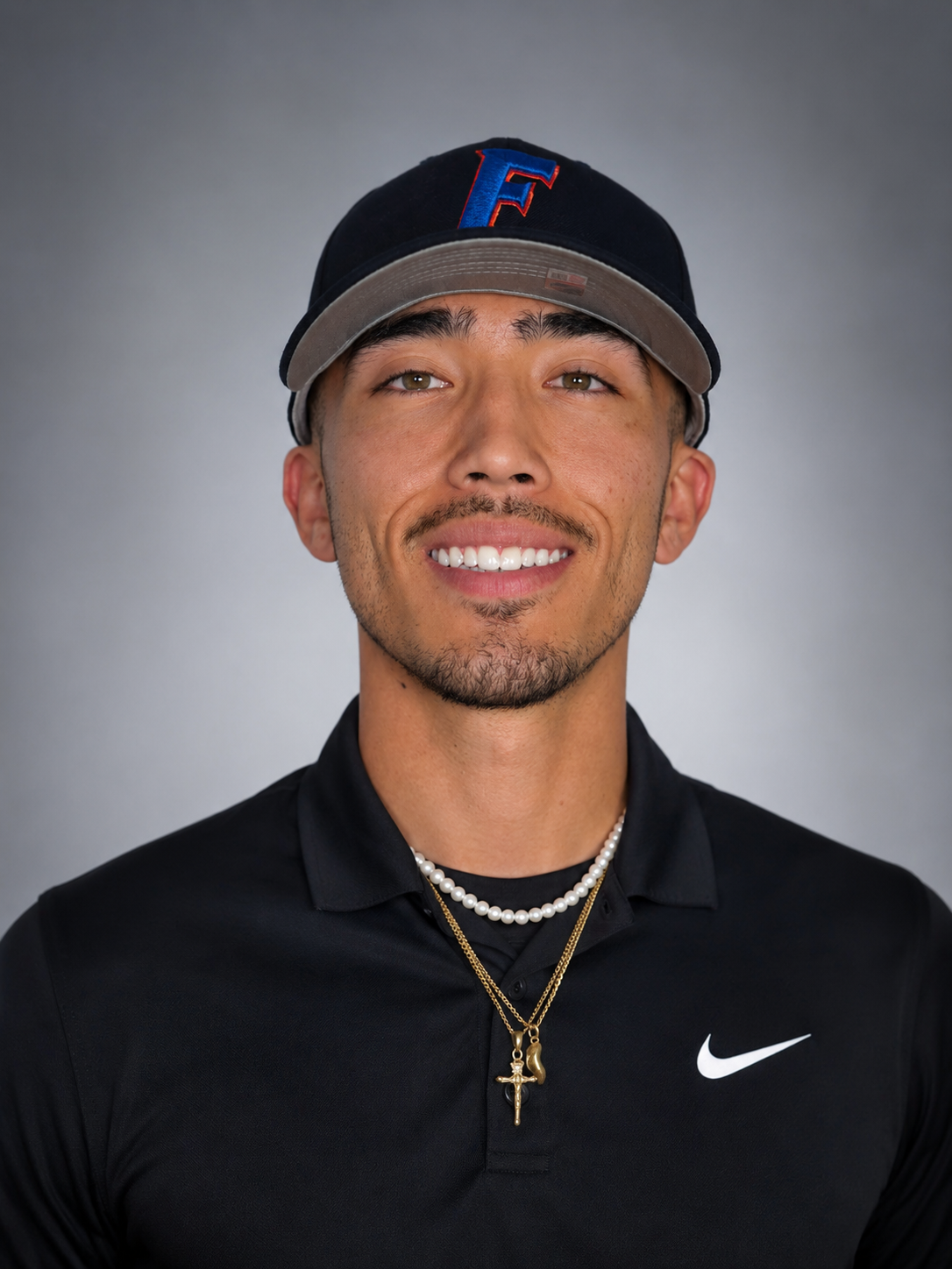 Close-up of a smiling young man wearing a dark baseball cap with a blue and red 'F' logo, a black Nike polo shirt, and layered necklaces including a pearl necklace and a gold chain with a cross pendant, against a plain gray background.