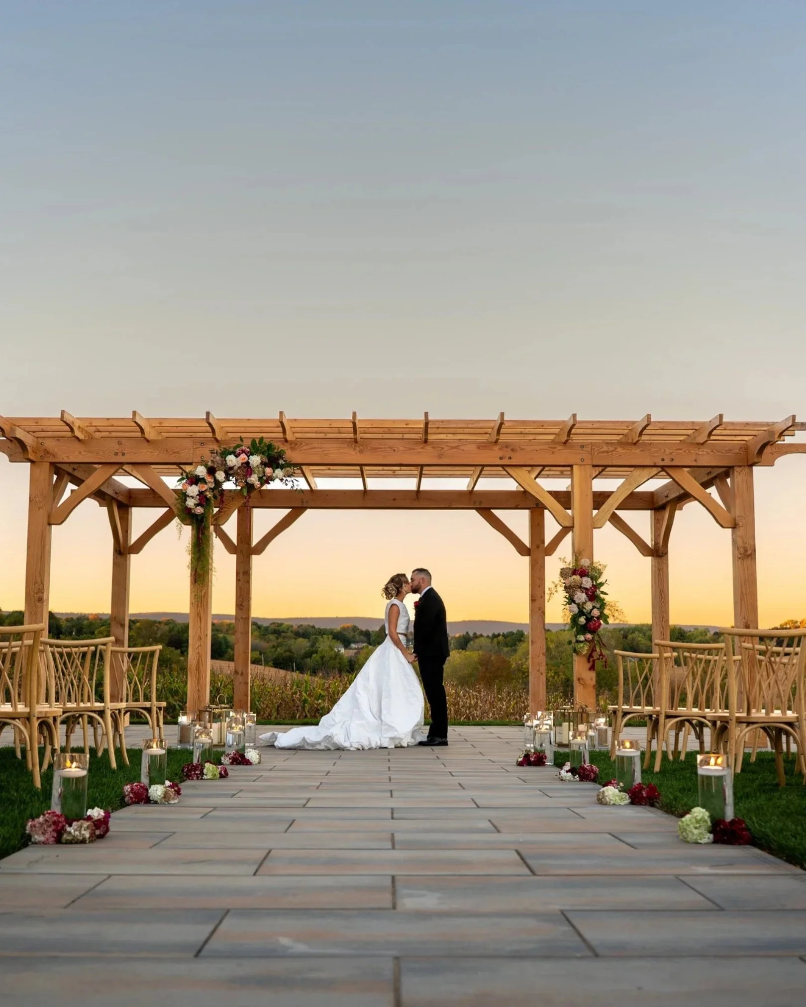 A bride and groom sharing a kiss under a pergola decorated with flowers during an outdoor sunset ceremony. Rows of chairs are on either side of the aisle, which is lined with floral arrangements and candles.
