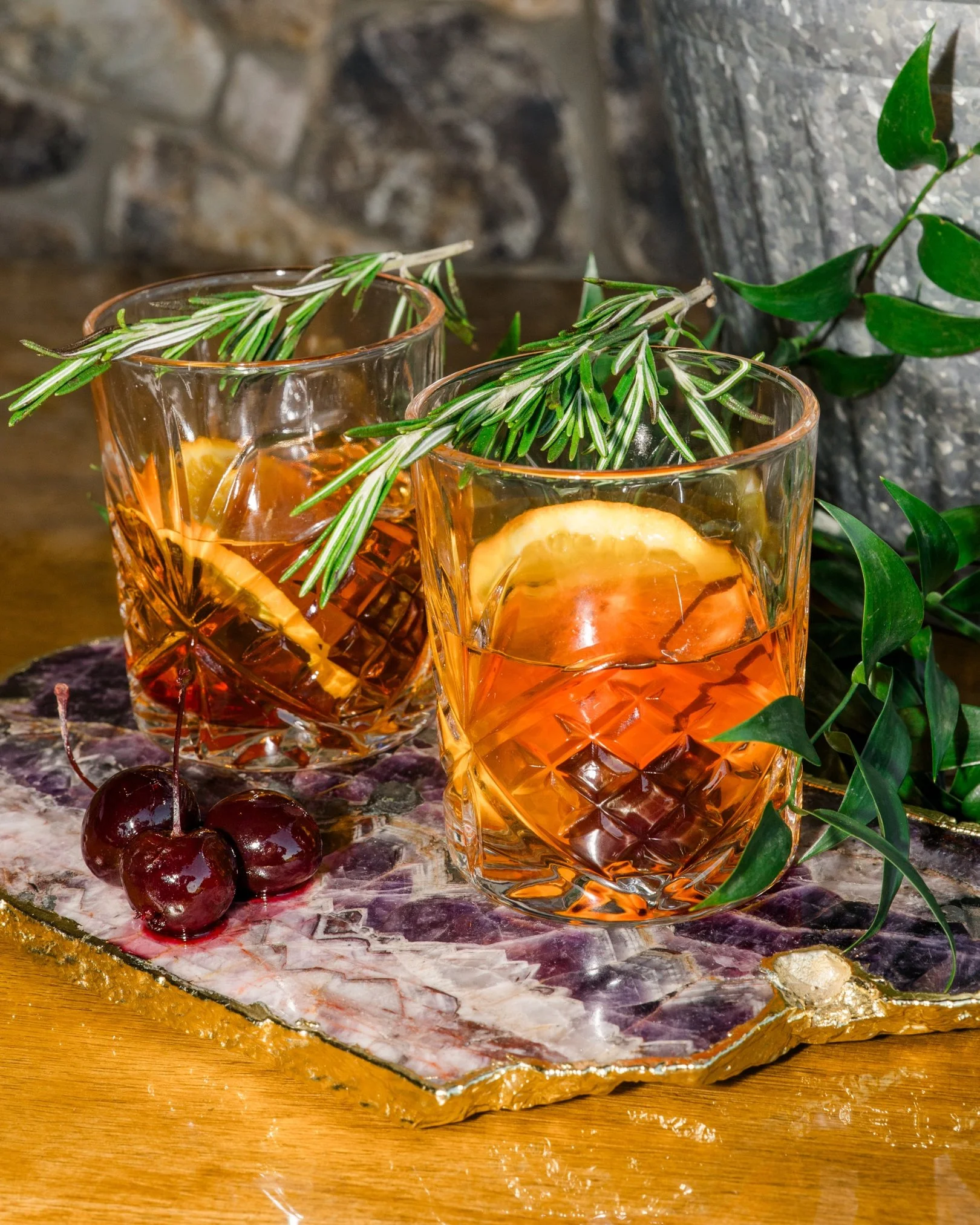 Two glasses of whisky garnished with orange slices and fresh rosemary sprigs, placed on a textured surface with cherries beside them, and green leafy plants in the background.